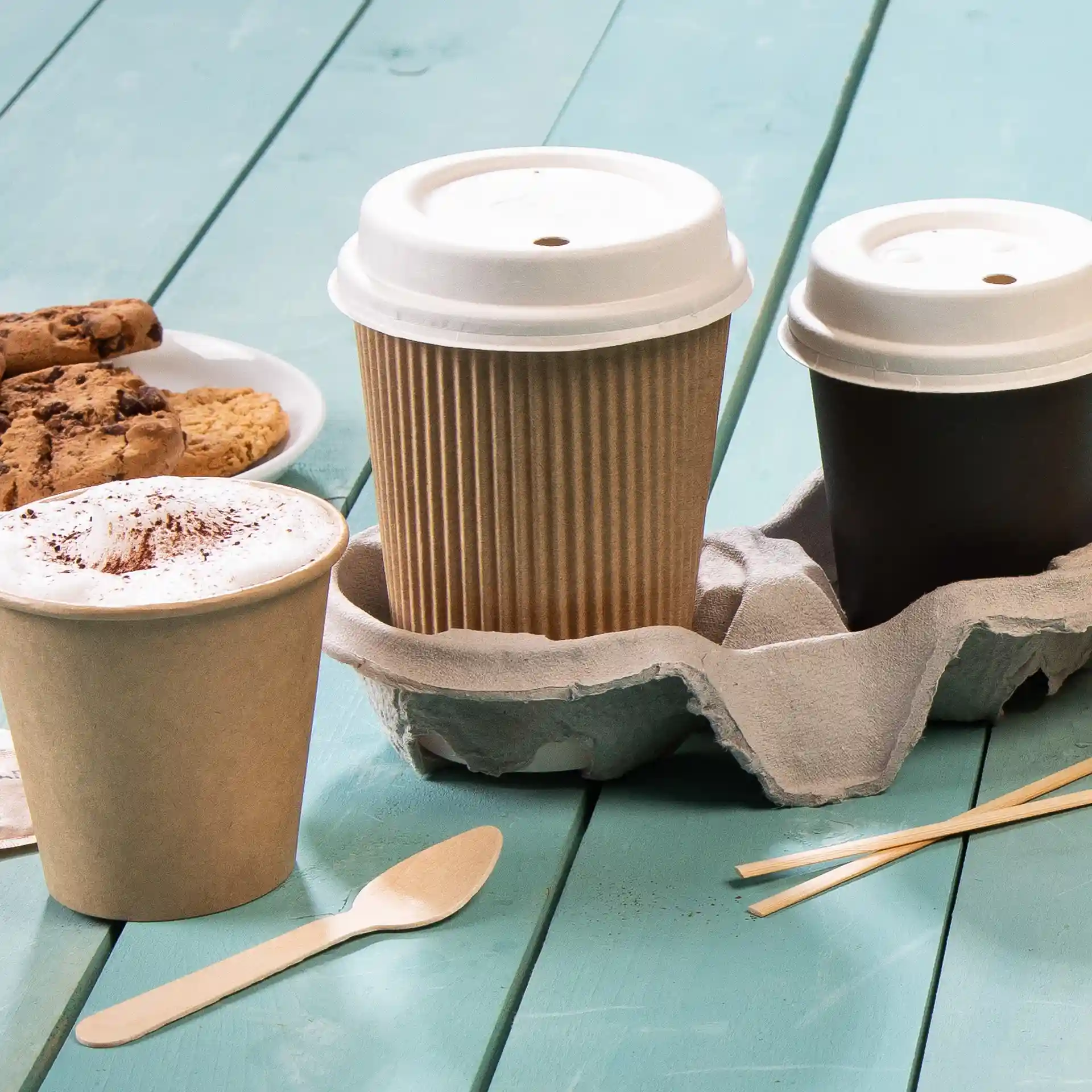 Image of a brown ribbed coffee cup, a white coffee cup, and a brown cup holder with a wooden spoon and cookies on a teal surface.
