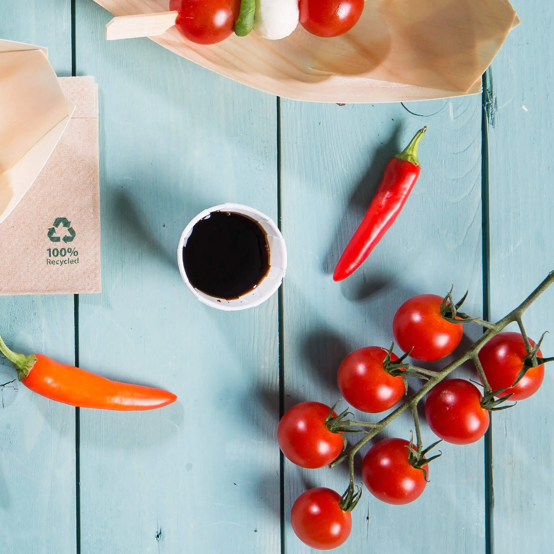 Image of a wooden plate with cherry tomatoes, chili peppers, a small white paper cup with dark sauce, and a brown wrapper with a recycling symbol.