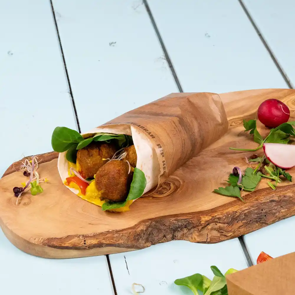 A wrapped snack filled with falafel, spinach, and sauce on a wooden board, accompanied by radishes and herbs.