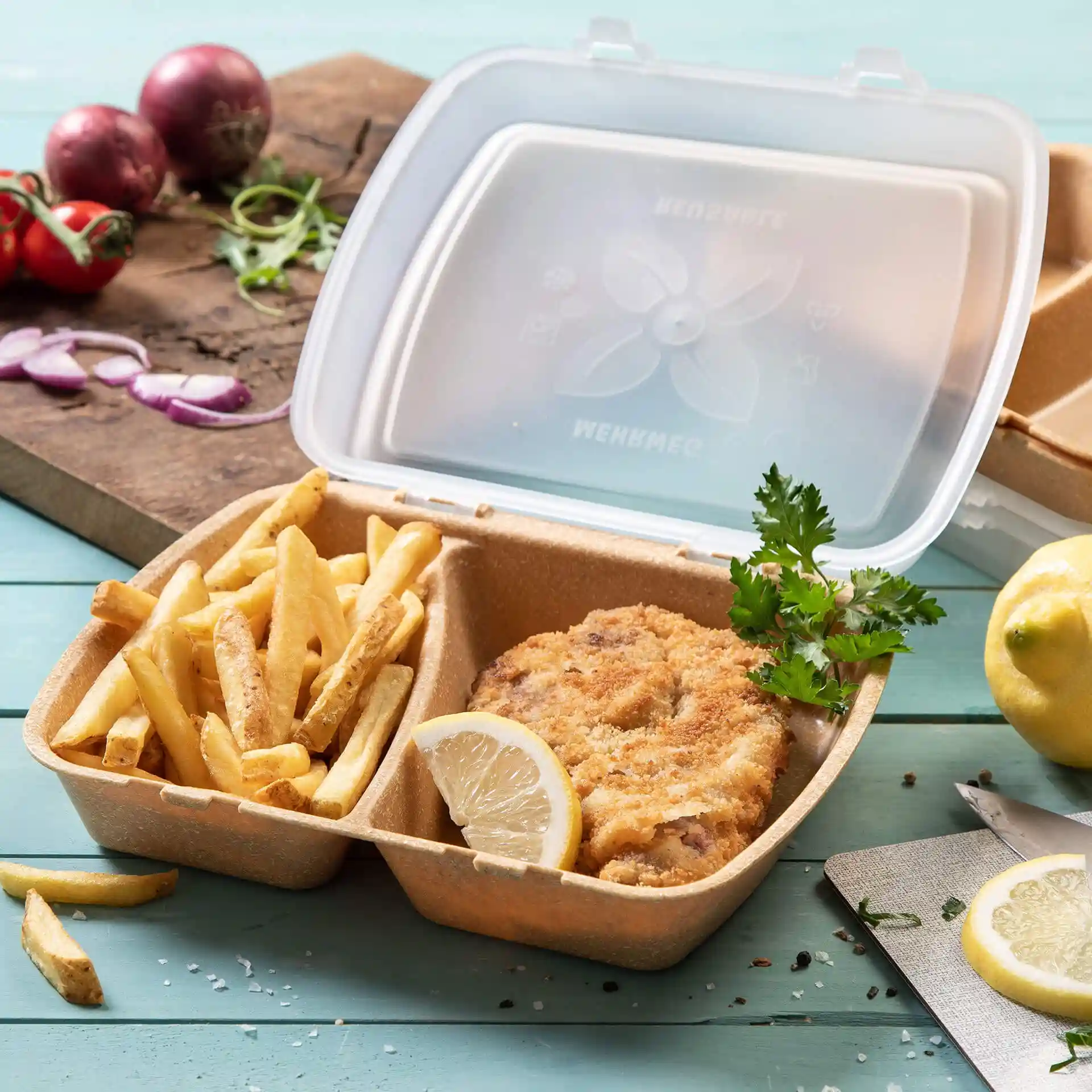 Container with fried chicken, fries, and lemon slice, inside a transparent plastic box on a blue surface.