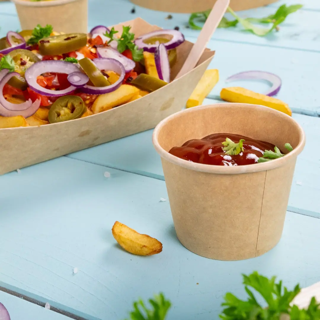 Brown paper cup filled with ketchup, garnished with a small piece of parsley, next to a serving of fries and vegetables.