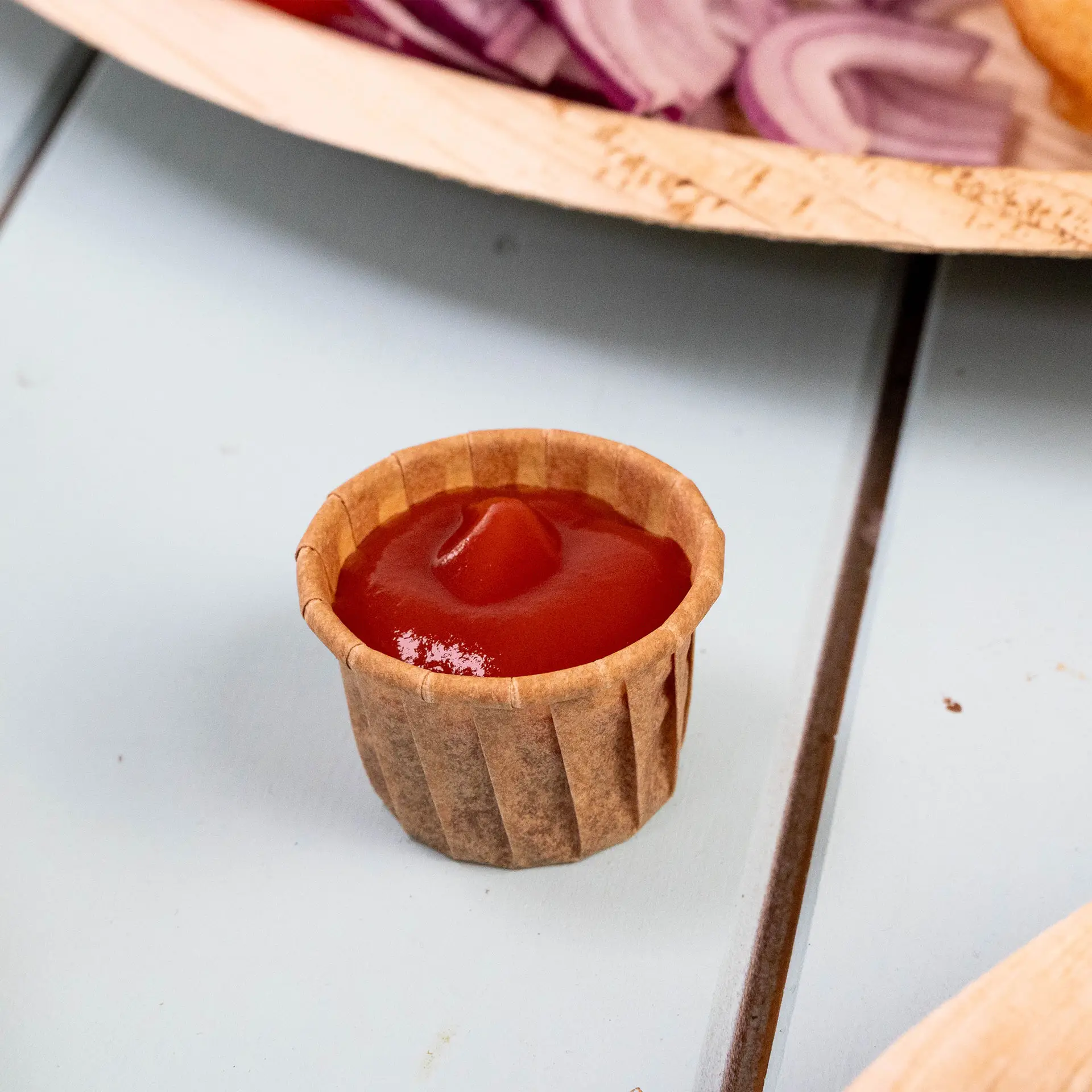 Brown 15 ml paper cup filled with ketchup, placed on a blue surface, with sliced onions and a plate in the background.