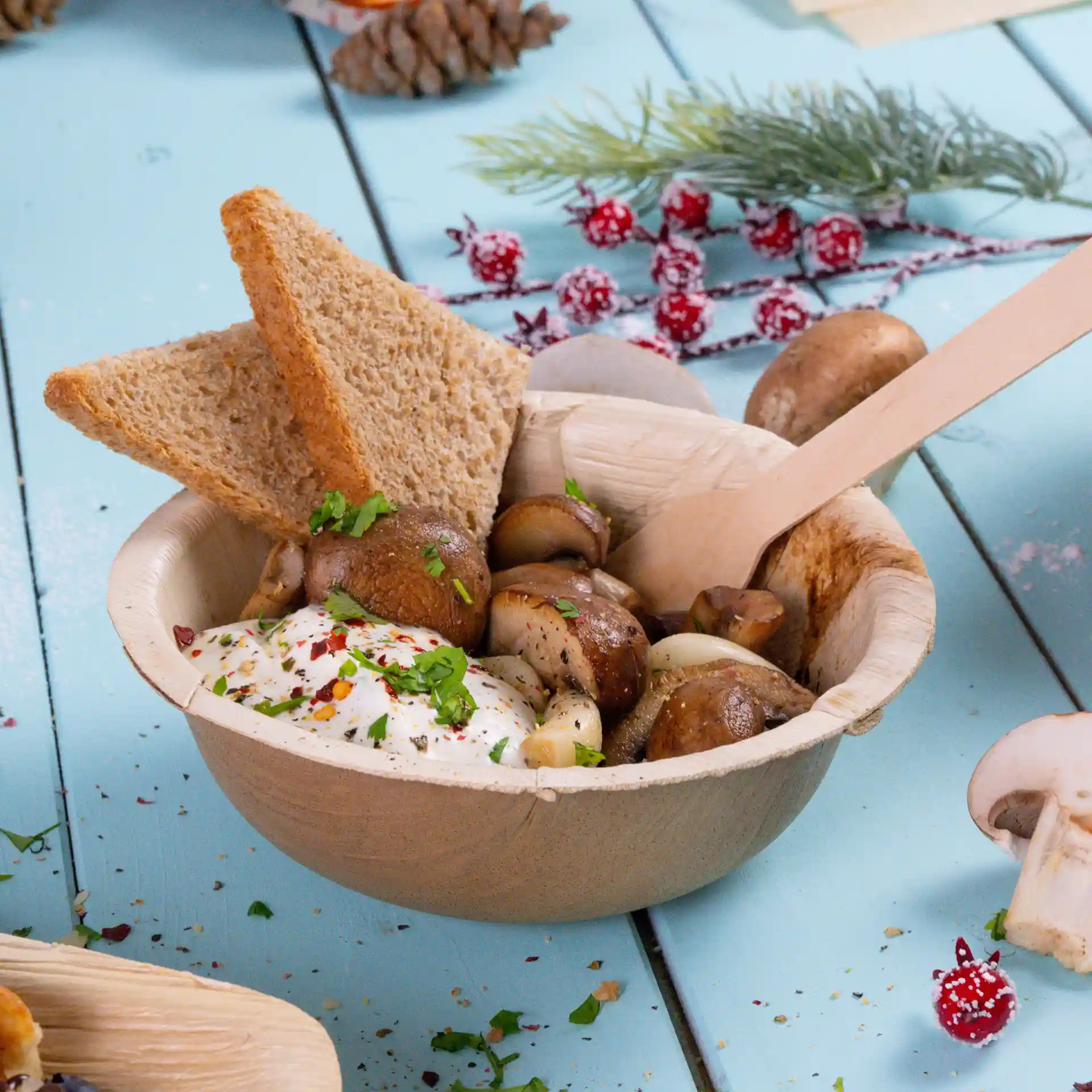 Bowl made of palm leaf containing mushrooms, a dollop of sauce, and two slices of brown bread, with a wooden spoon.