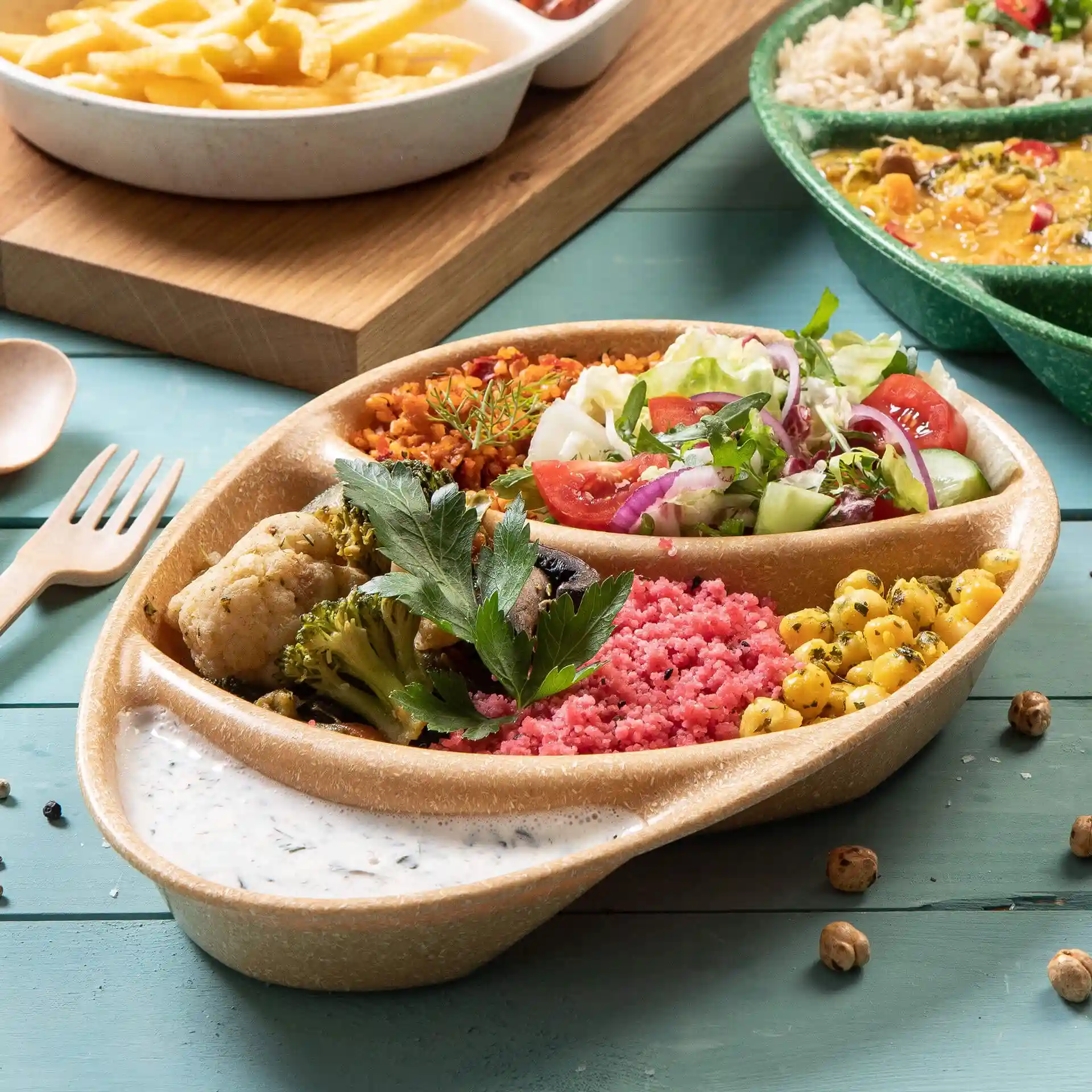 Brown reusable plate with three compartments, filled with various foods and a sauce on a blue wooden surface.
