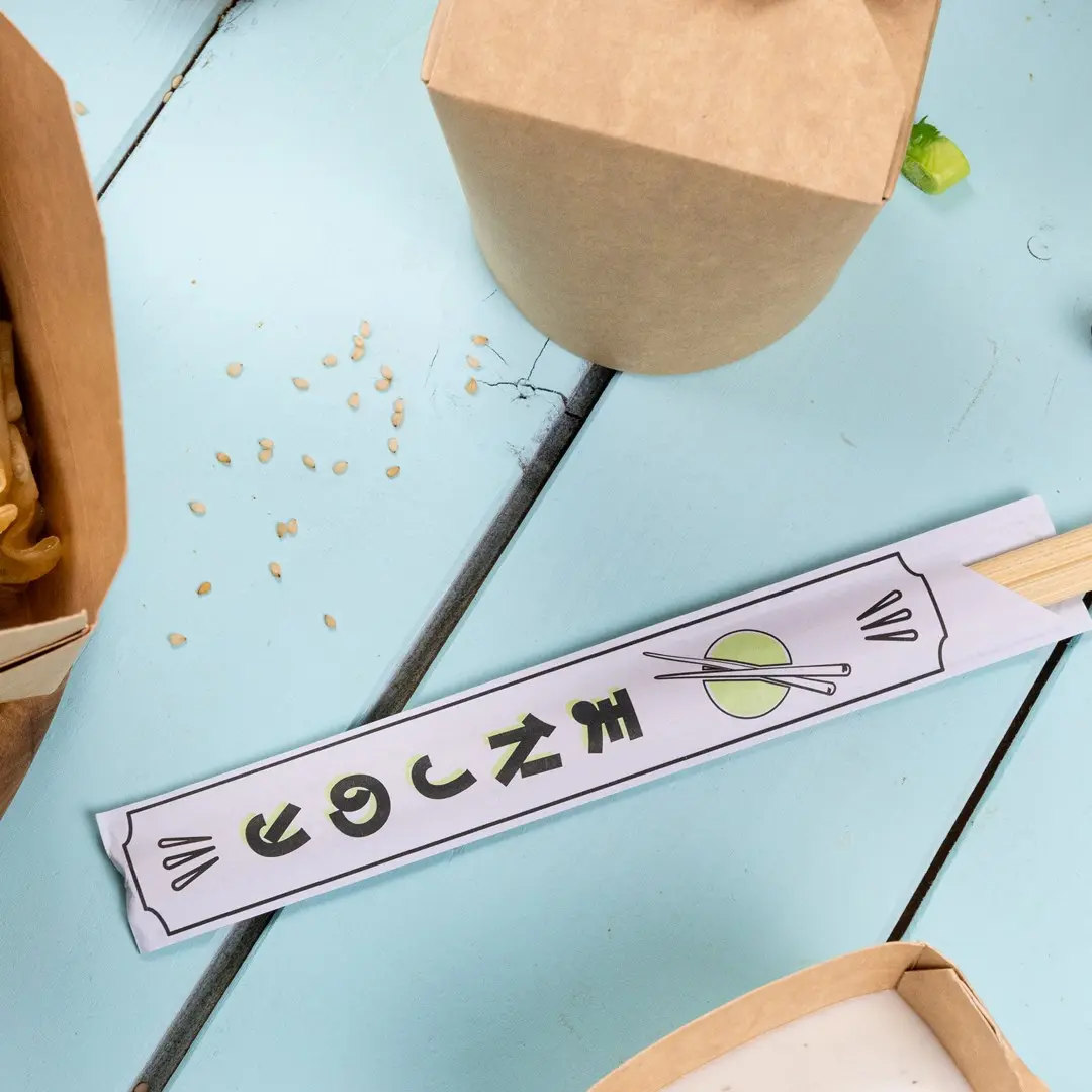 Chopstick wrapper with graphic design and chopsticks on a blue wooden surface, surrounded by food containers and sesame seeds.