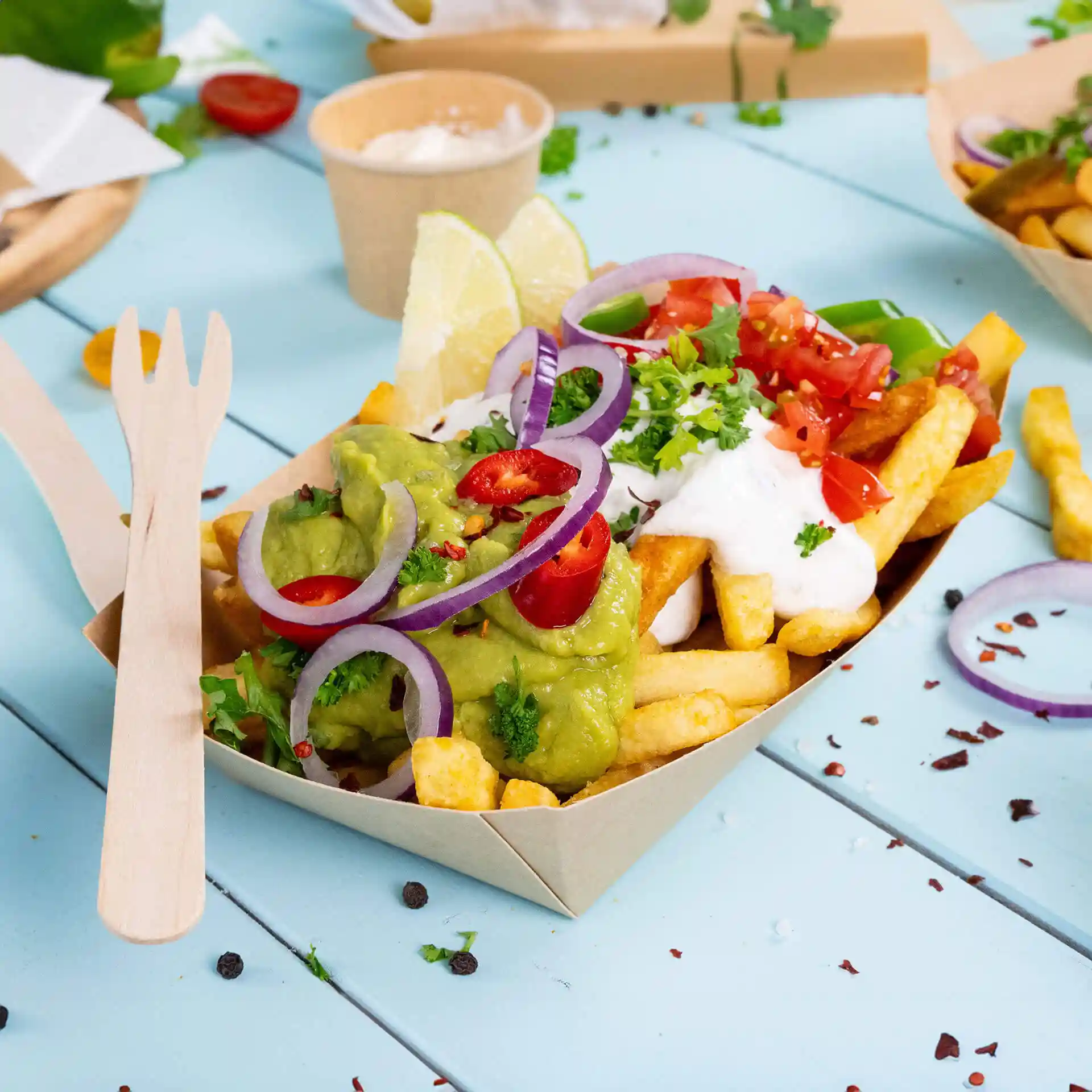 Deep cardboard snack bowl filled with fries, guacamole, sour cream, diced tomatoes, and garnished with onions and lime.