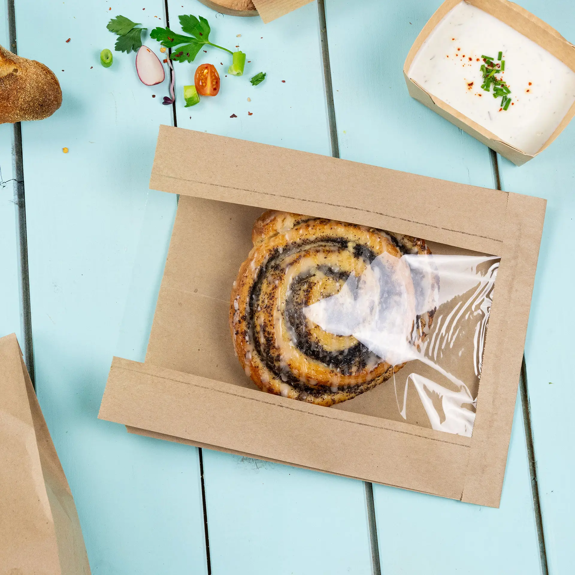 One brown flat paper bread bag with a transparent PLA window showcasing a swirl pastry inside; additional garnishes and condiments nearby.