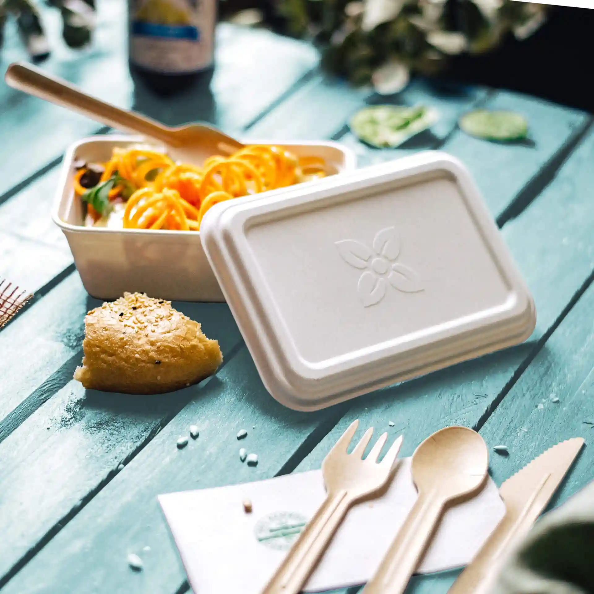 Rectangular white sugarcane container with a lid, fork, spoon, and a piece of bread on a blue wooden surface.