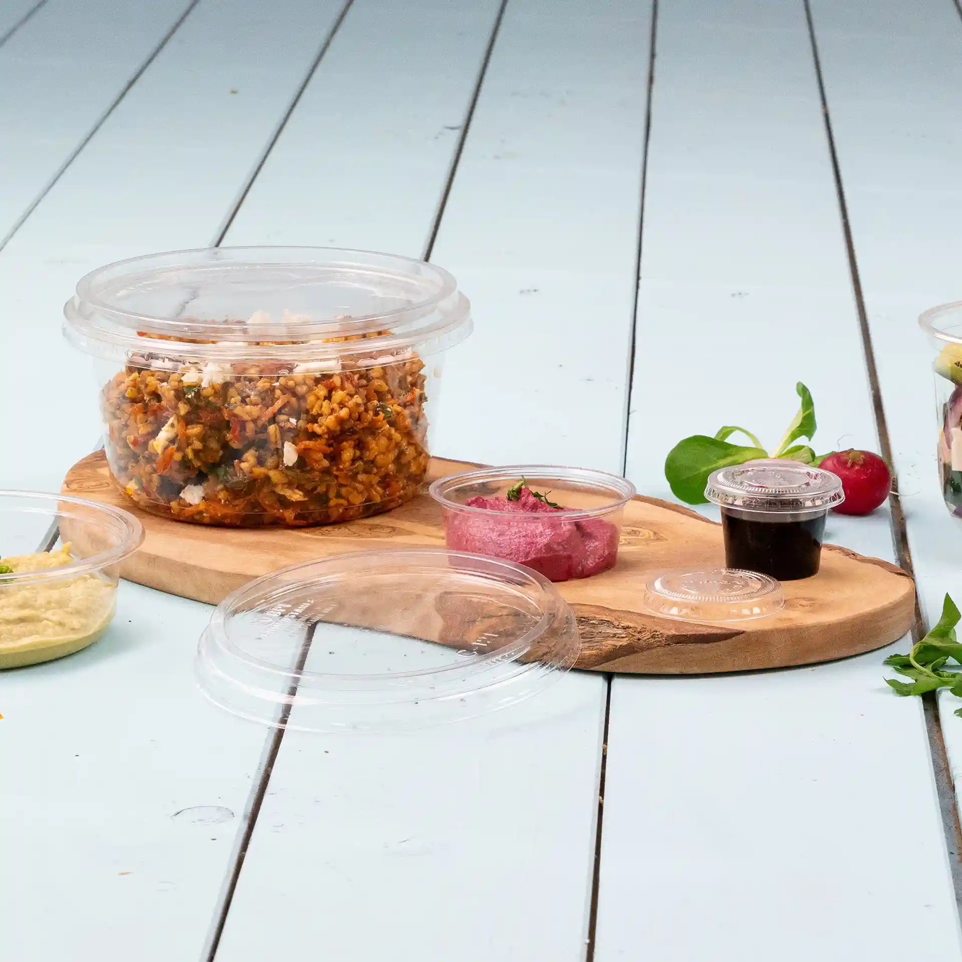 Transparent round deli container with a lid, filled with food, on a wooden board with small sauce cups nearby.