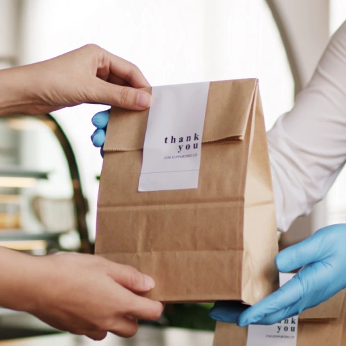 Two hands exchanging a brown paper bag with a "thank you" label on it, in a café setting.