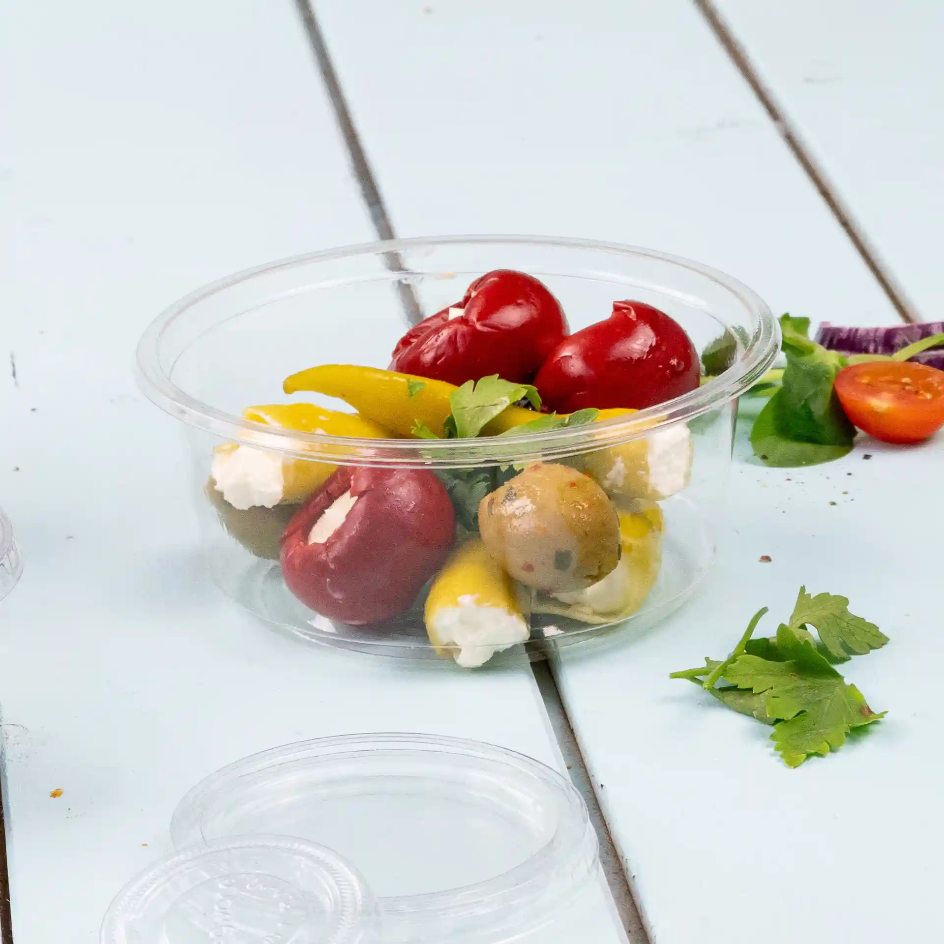 Transparent round deli pot containing assorted peppers, olives, and cheese, placed on a light blue wooden surface.