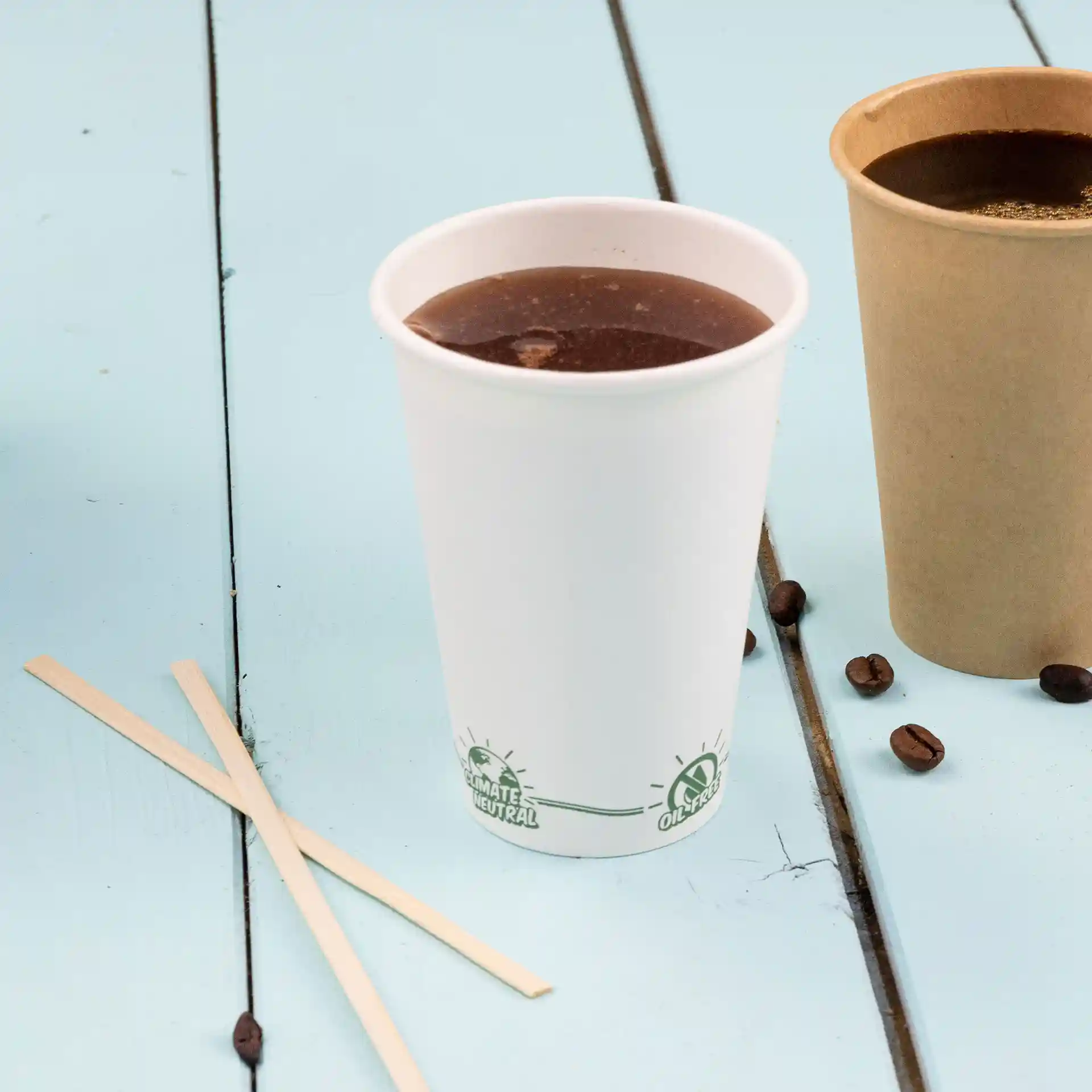White paper cup filled with dark liquid, next to a brown paper cup and bamboo stirrers on a blue wooden surface.