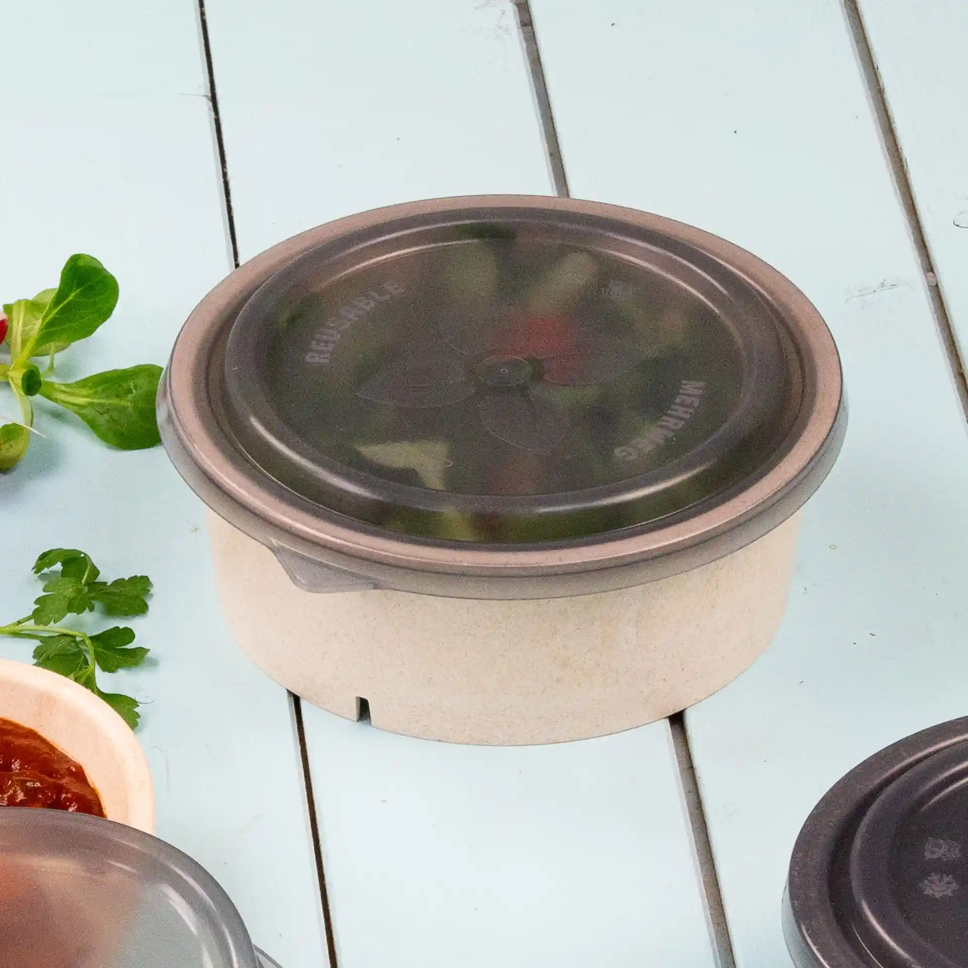 Round grey bowl with a grey lid, made of PP, placed on a light blue surface with some herbs nearby.