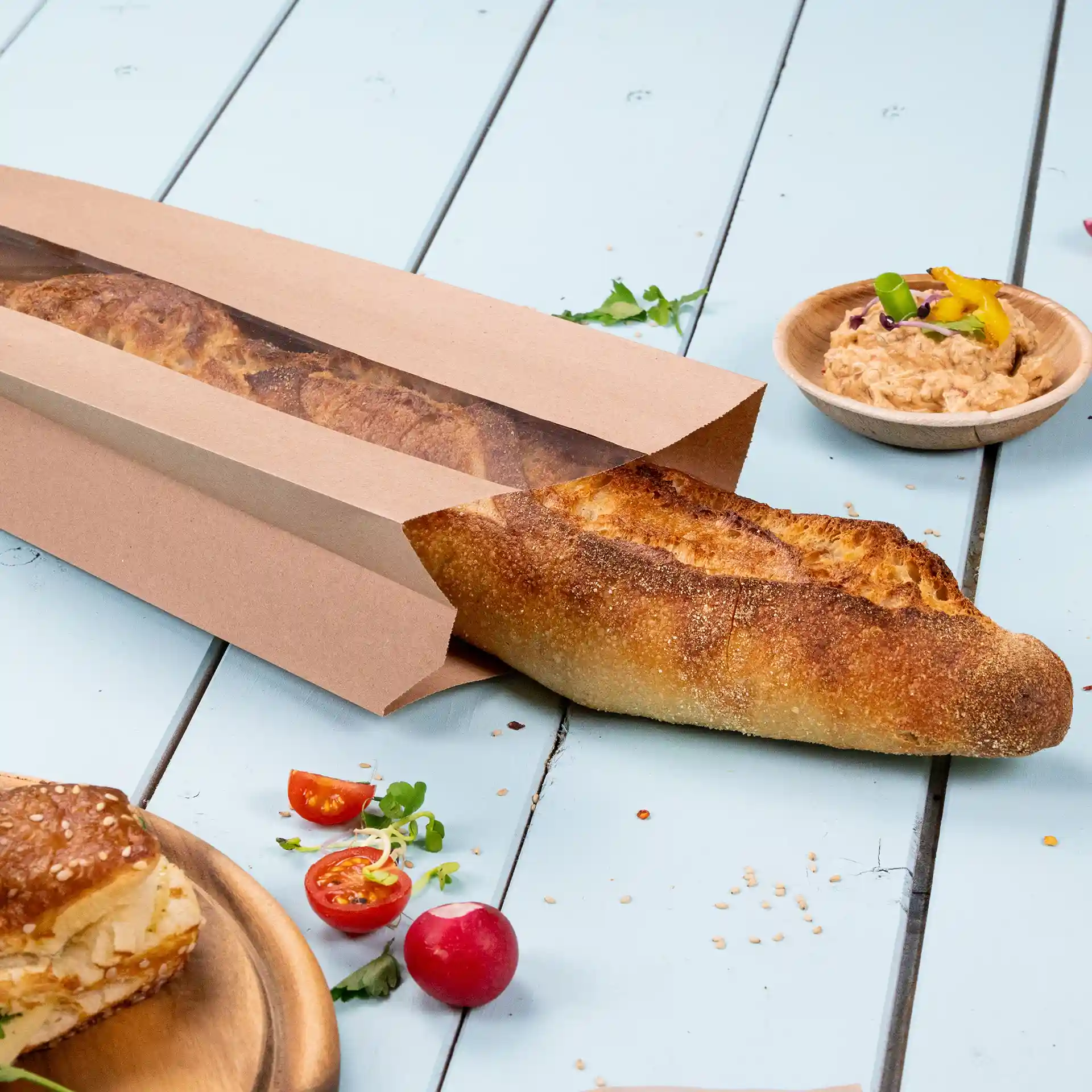 Brown flat paper bread bag with a window, containing a baguette, placed on a light blue surface with tomatoes and dip nearby.