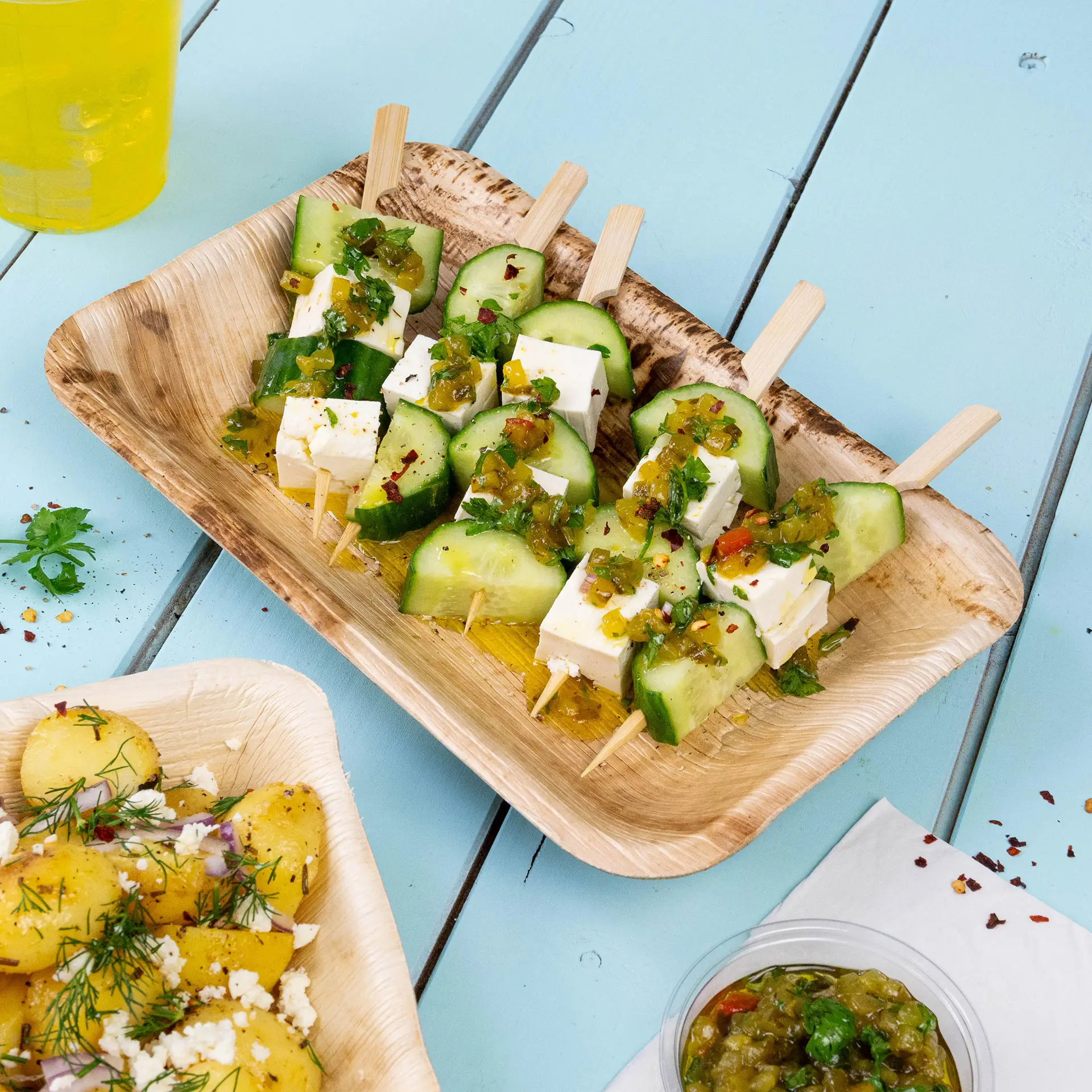 Bamboo plate with cucumber and cheese skewers topped with herbs and sauce, served with a side of potato salad.