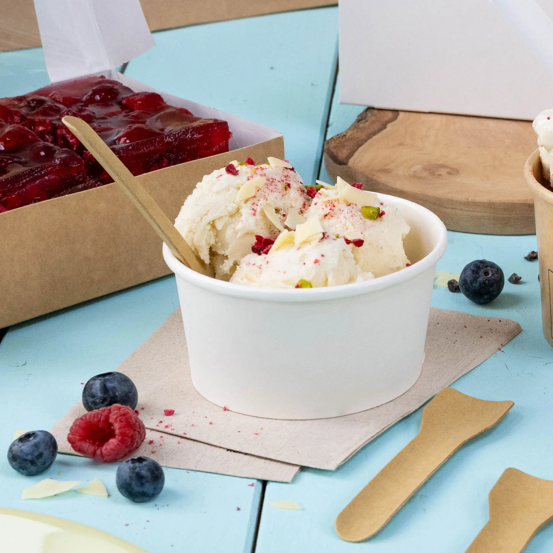 Cup of ice cream topped with fruit and nuts, wooden spoon, and fresh berries on a blue surface with a napkin.
