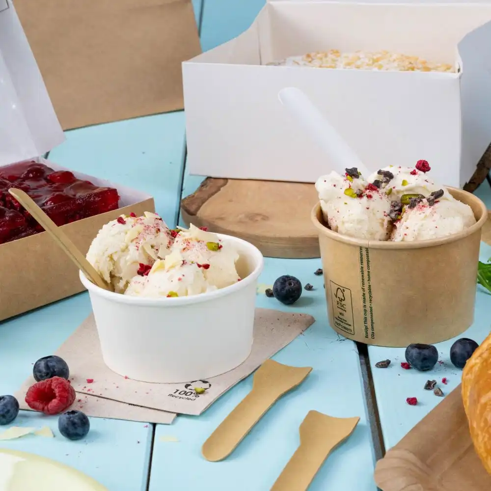 Two ice cream cups on a light blue table: one white with toppings, the other brown with chocolate and berry toppings. Surrounding items include wooden spoons and various fruits.