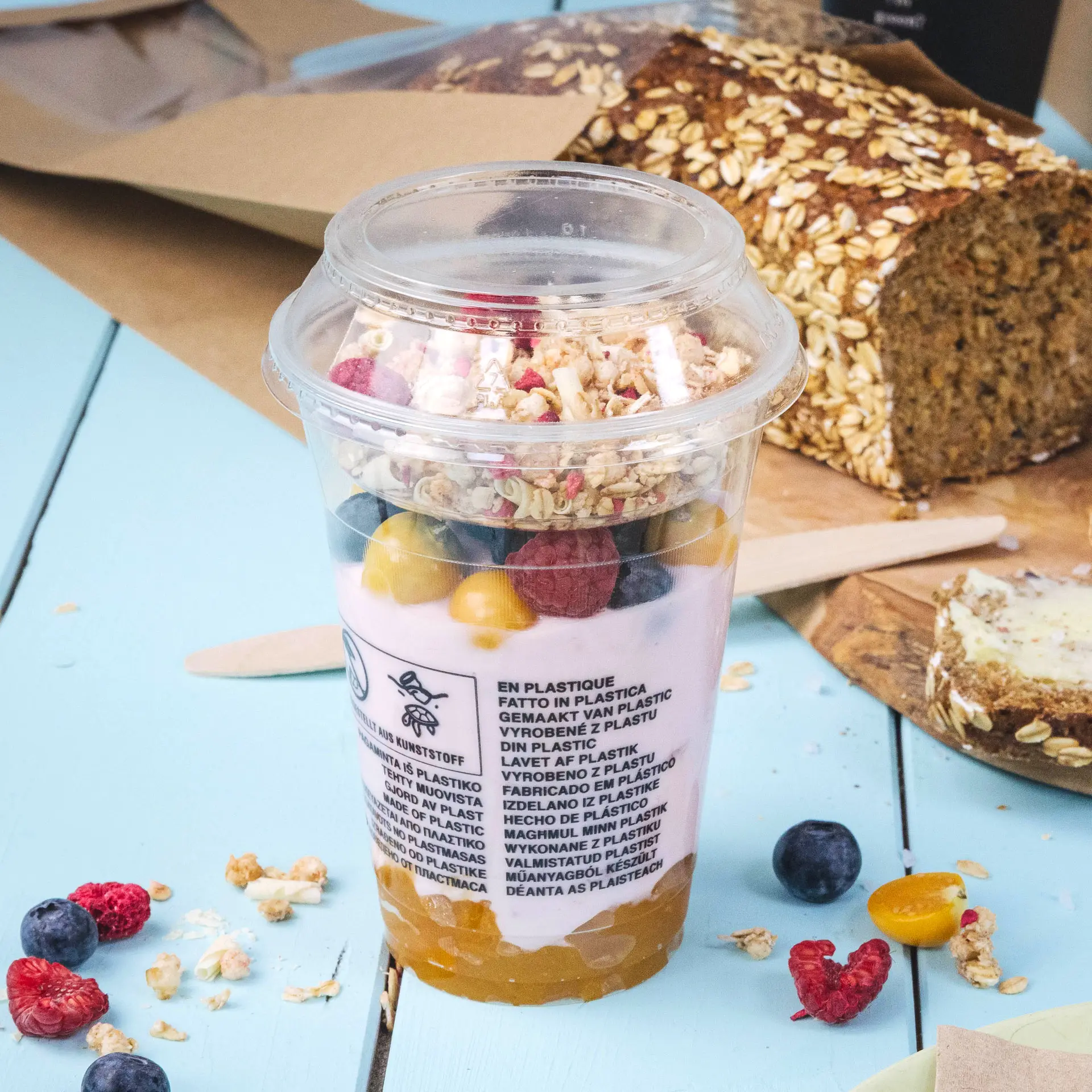 A clear 300 ml PLA smoothie cup filled with yogurt, fruits, and granola, placed on a light blue table with a loaf of bread nearby.