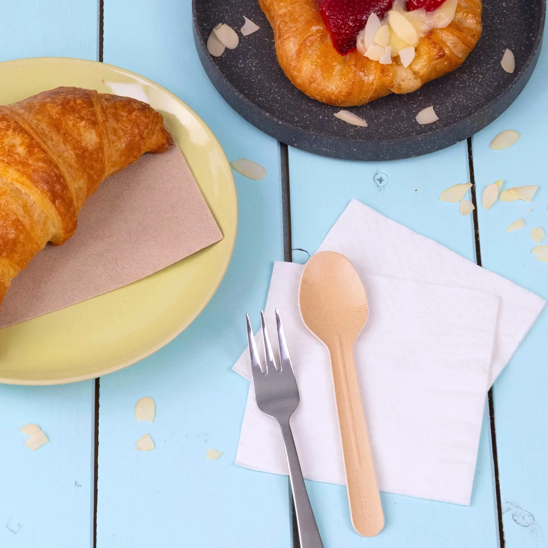 Image of a croissant on a yellow plate, a pastry on a dark plate, and a paper spoon next to a fork on napkins.