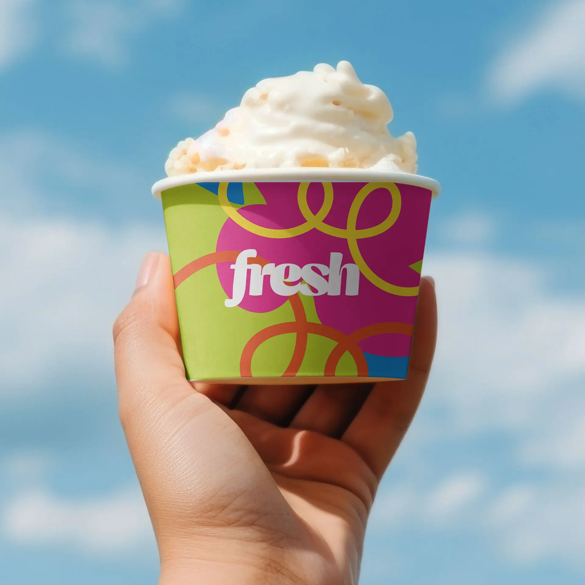 A hand holding a colorful ice cream cup with a fresh design and whipped cream on top against a blue sky.
