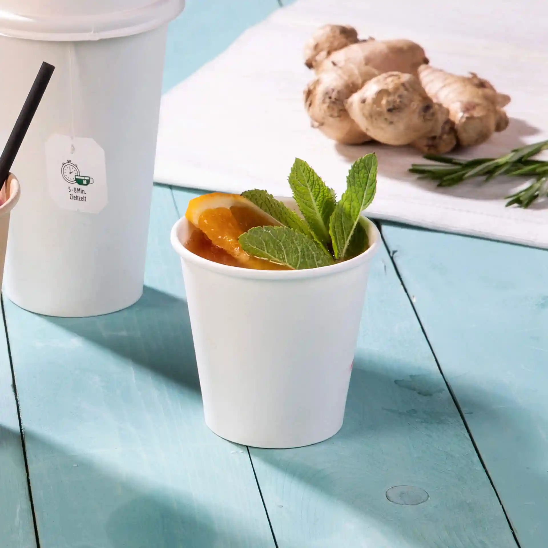 White paper cup with a drink, garnished with mint leaves and orange slices, on a blue wooden surface.