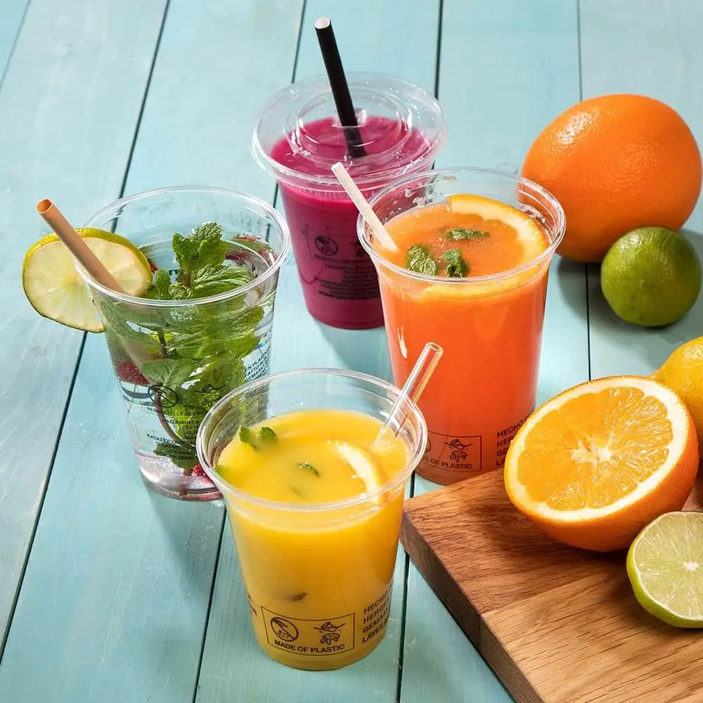 Four clear plastic cups filled with colorful drinks, including orange juice and a pink smoothie, beside fresh fruits.