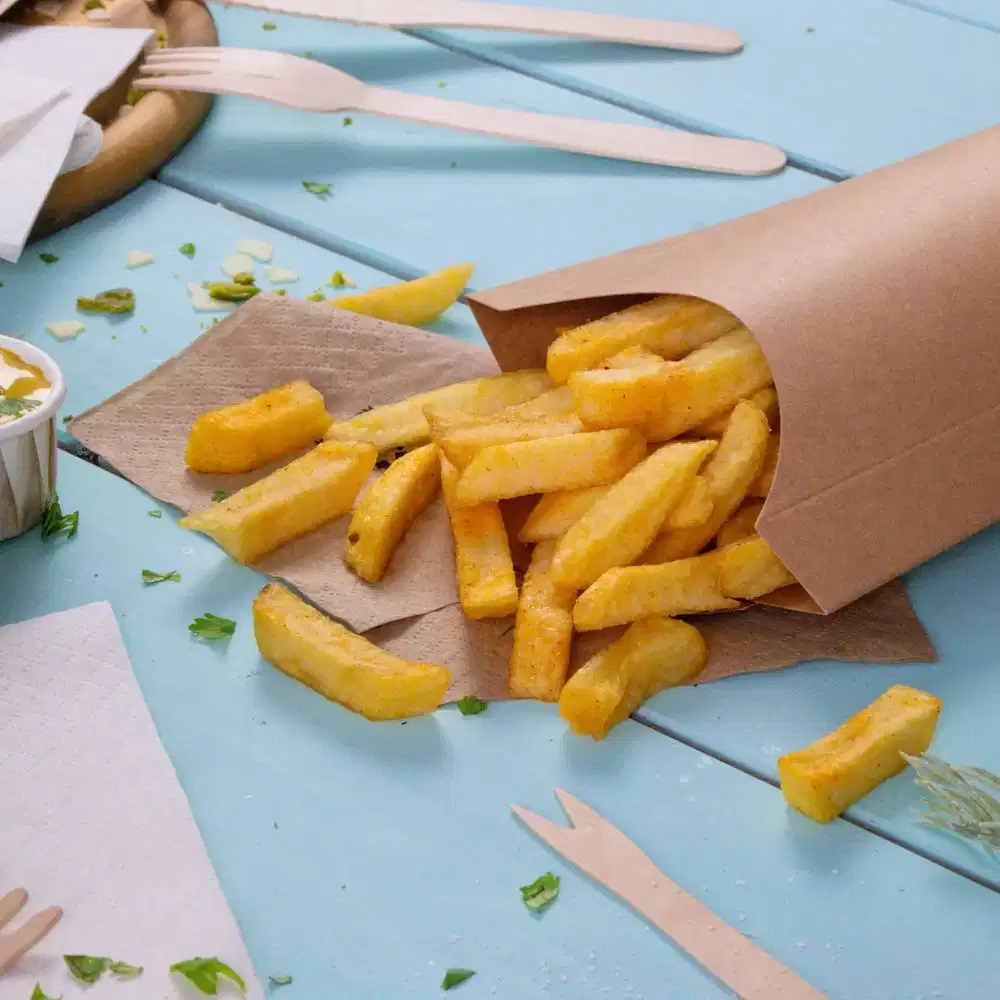 Brown paper container spilling golden French fries on a blue table with napkins and wooden utensils nearby.