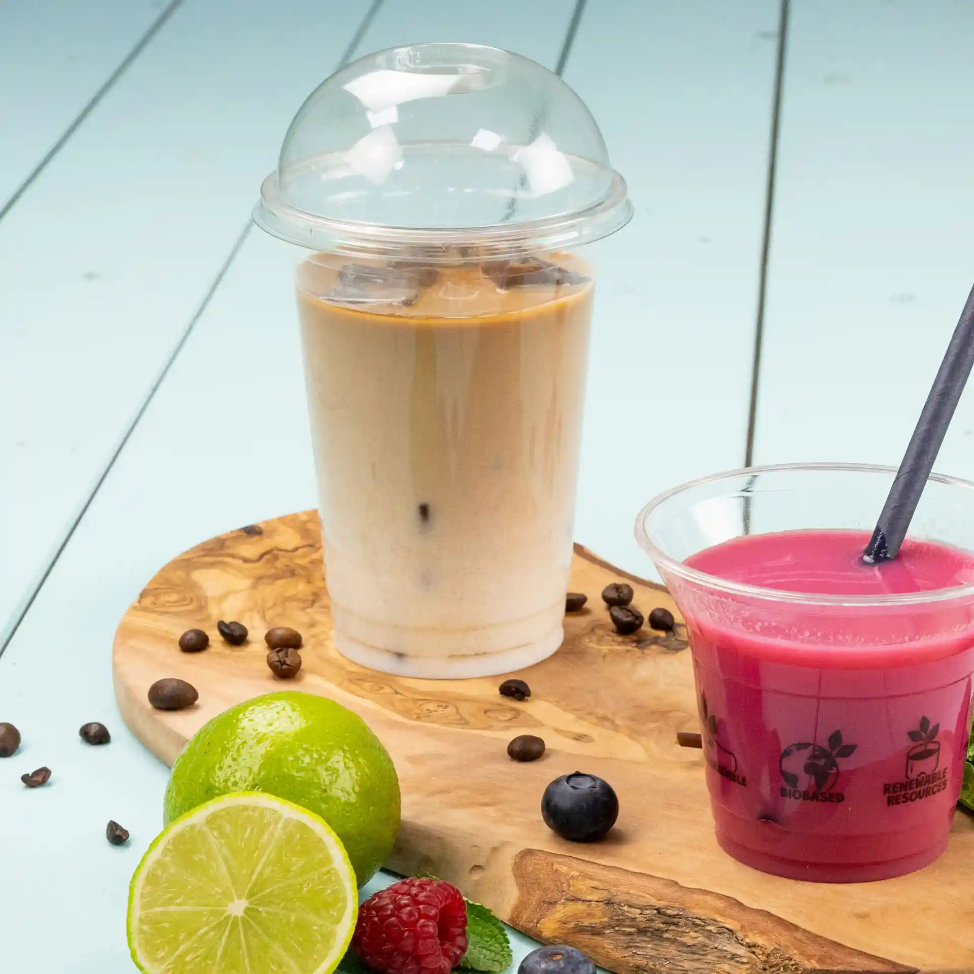 Image of a transparent 16 oz plastic cup filled with iced coffee, next to a pink beverage in a clear cup on a wooden board with fruits.