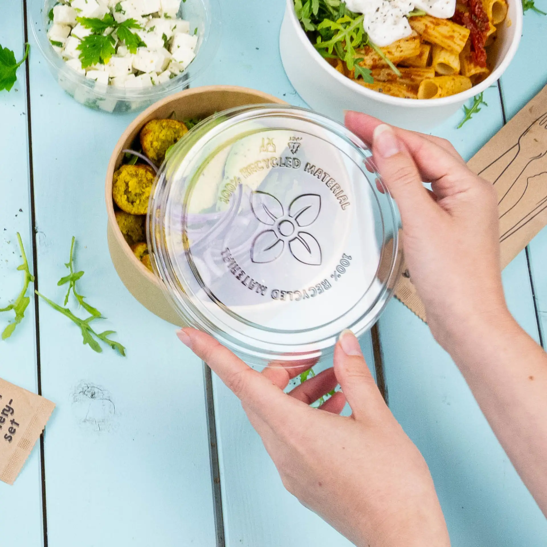 Clear rPET lid for a PLA deli container being held above a bowl of food; background includes fresh ingredients.