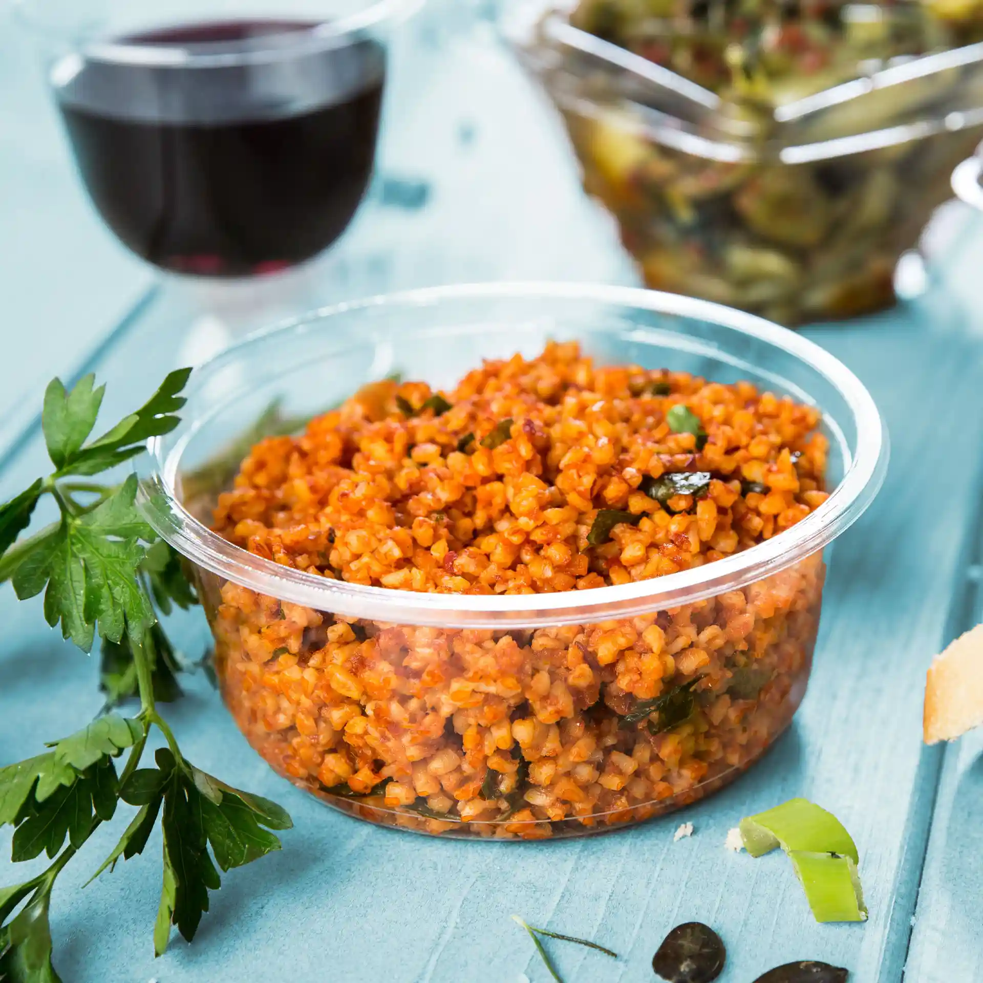 Round transparent deli container filled with orange bulgur, garnished with parsley, on a blue surface.