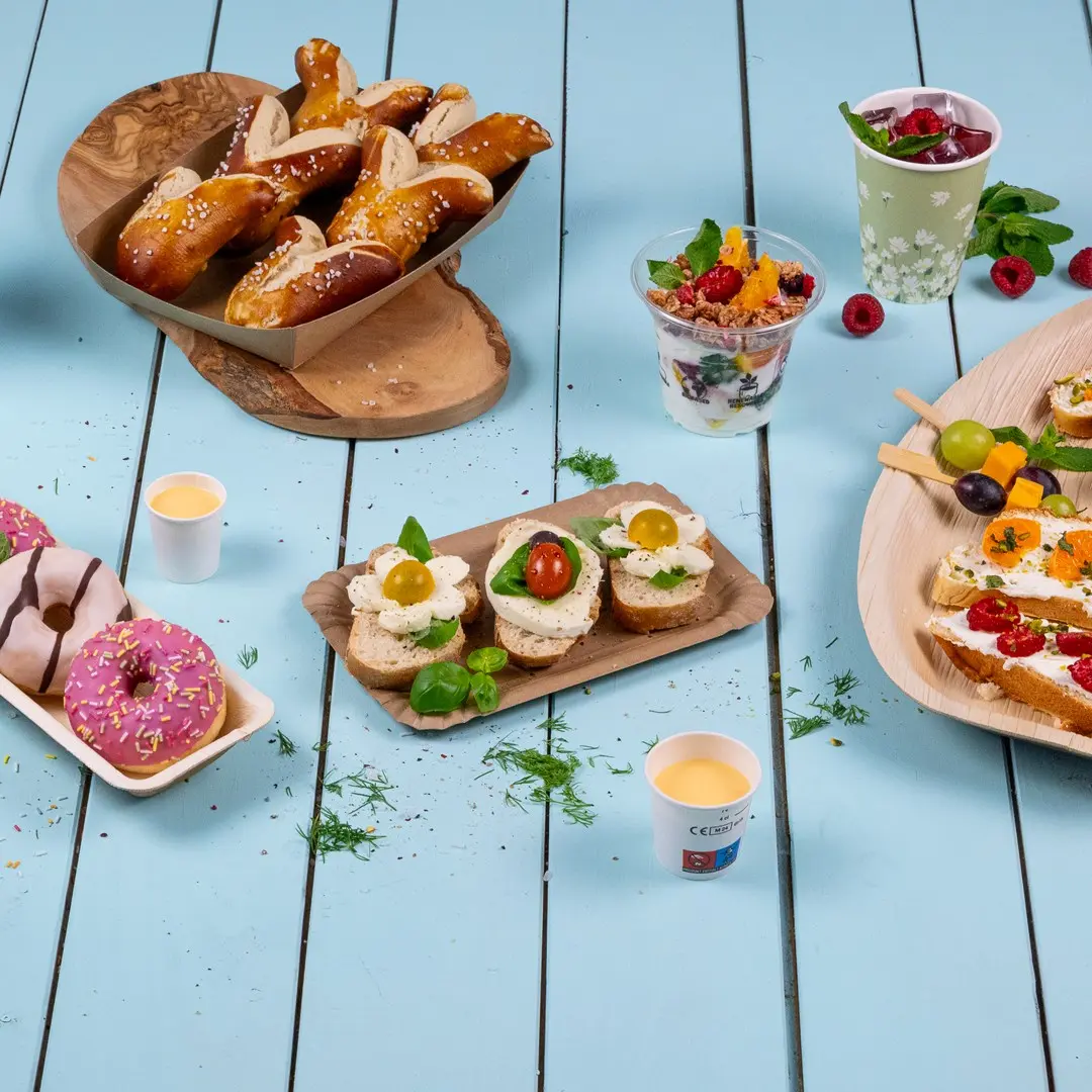 A variety of finger foods including pretzels, sandwiches with toppings, donuts, and fruit cups on a blue wooden table.
