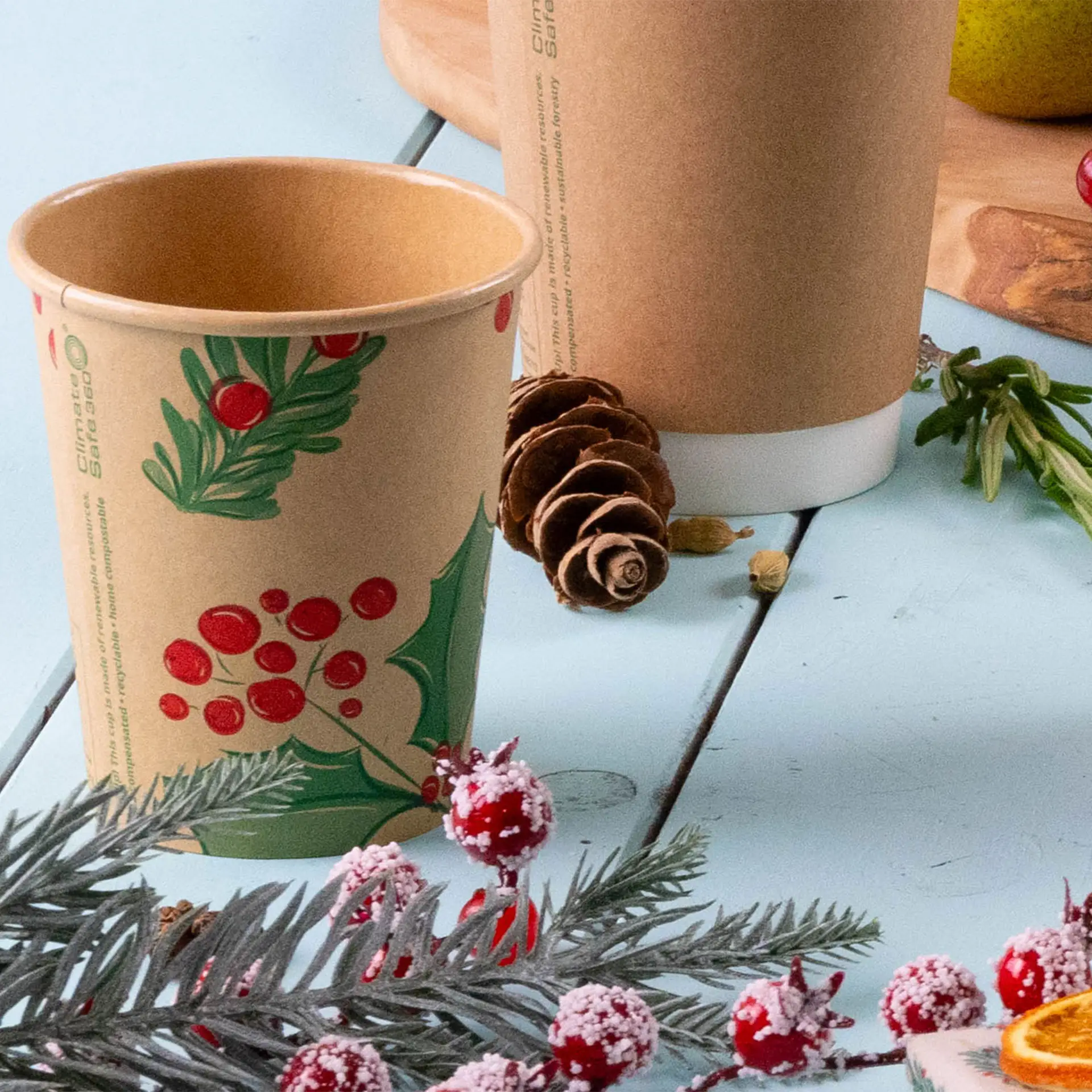 Brown paper cup with winter-themed design featuring red berries and green leaves, surrounded by pinecones and decorative elements.