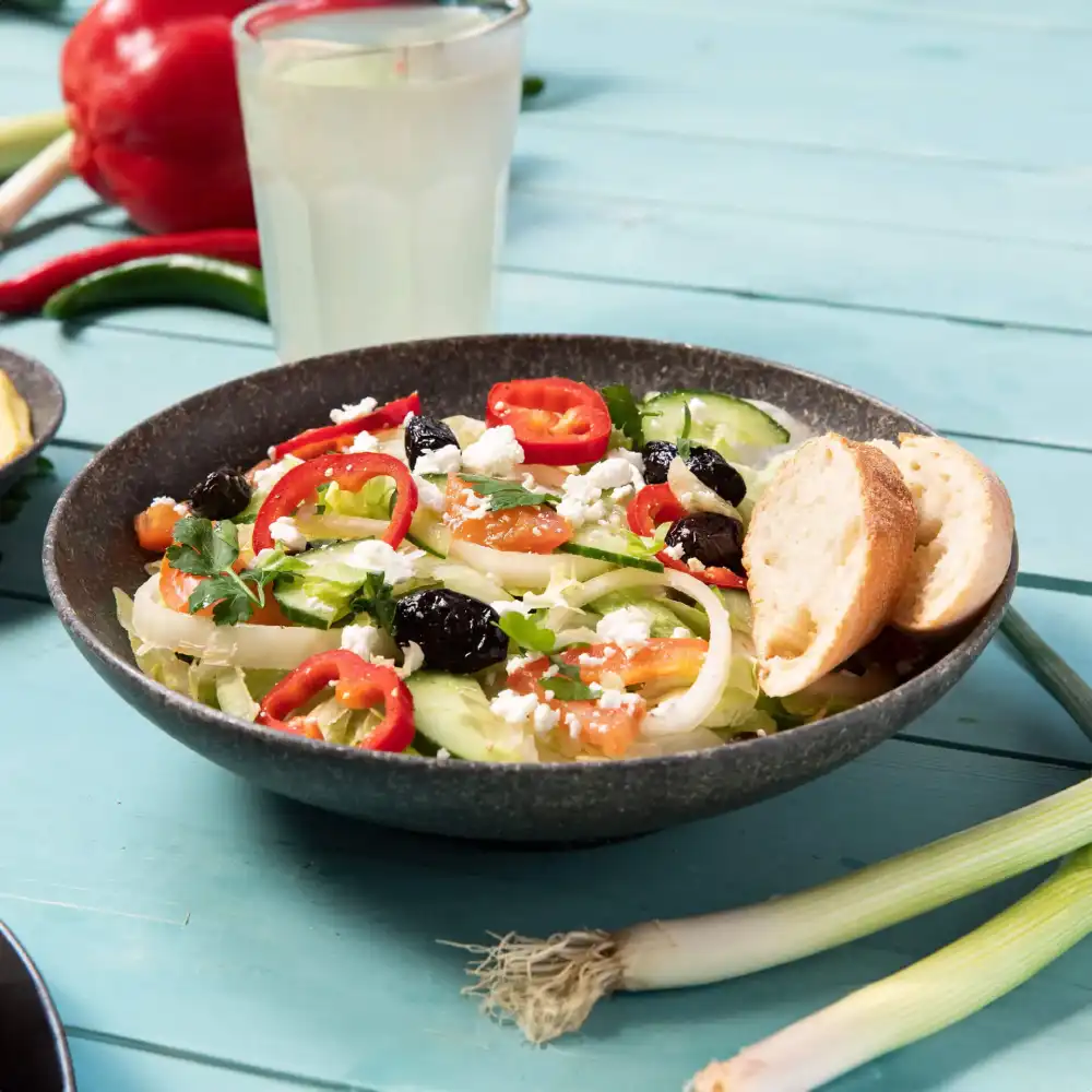 Bowl of salad with cucumber, tomatoes, olives, feta cheese, and red pepper slices, accompanied by two pieces of bread.