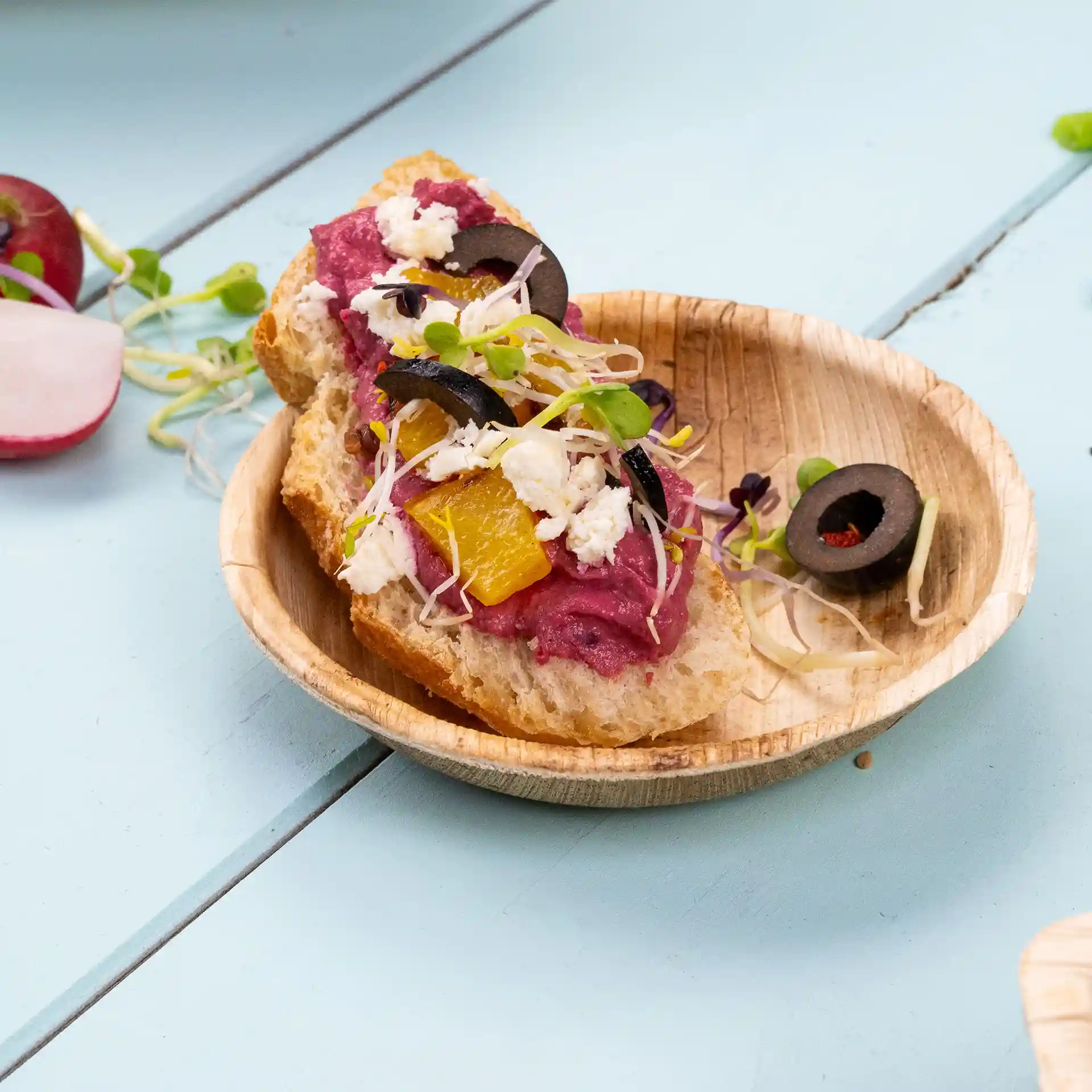 Round palm leaf bowl containing a slice of bread topped with purple spread, olives, sprouts, and orange pieces.