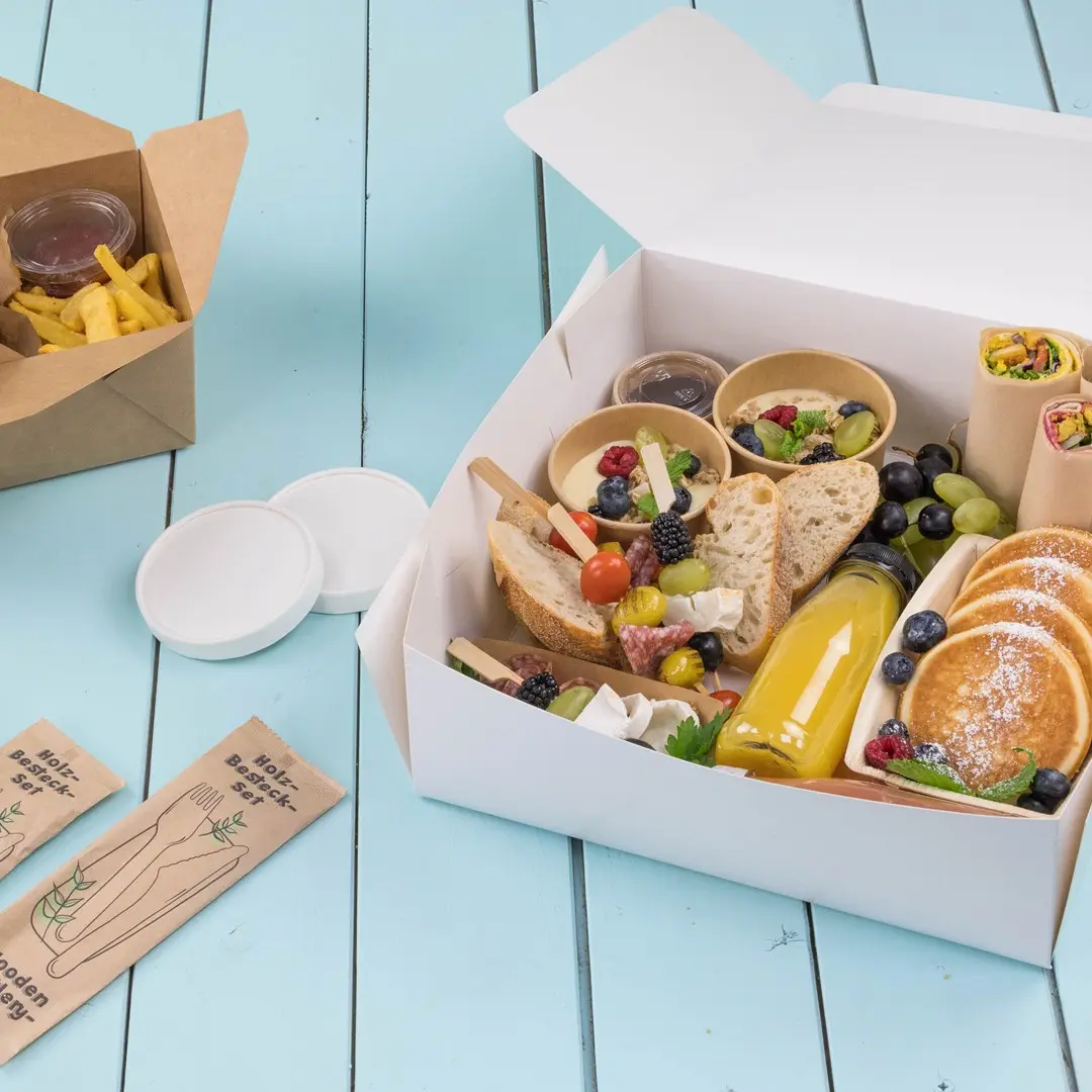 White take-away box filled with assorted food items, including bread, fruits, pancakes, and a drink, on a blue wooden surface.