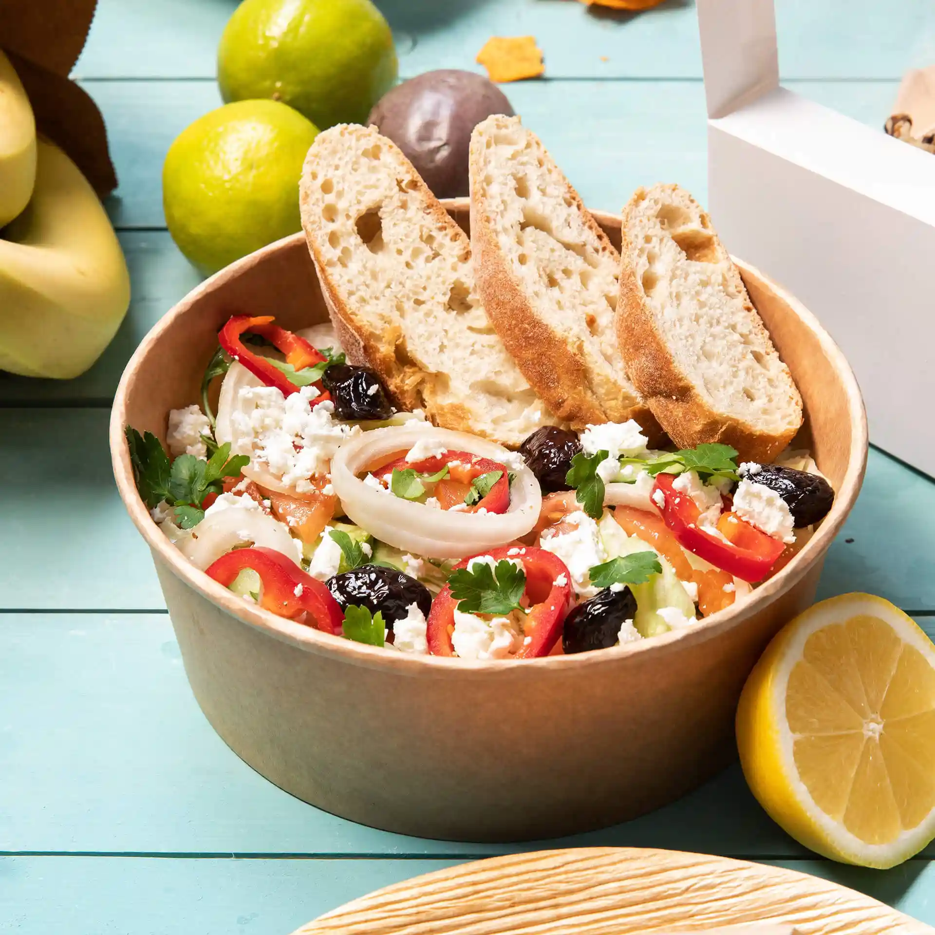 Round brown paper bowl filled with salad, topped with sliced bread, surrounded by fruits and a white takeout box.