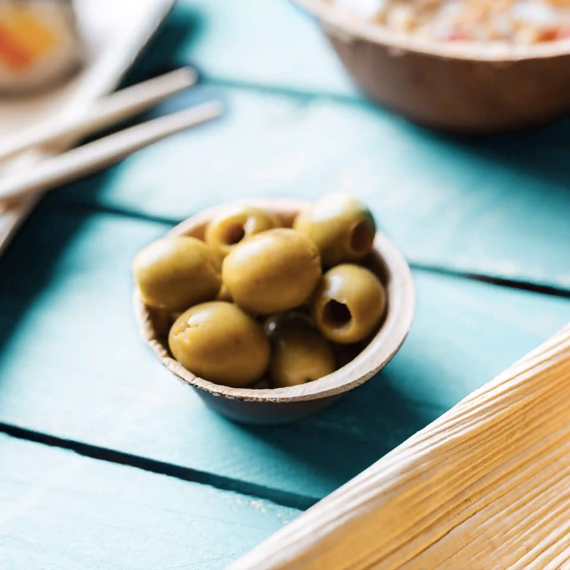 Bowl made of palm leaf containing several green olives on a turquoise wooden surface.