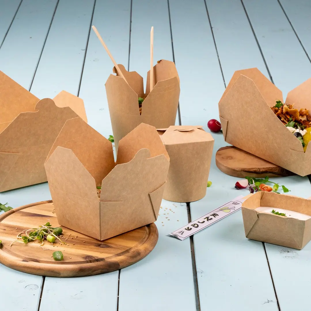 Brown cardboard food containers in various shapes on a wooden board with fresh ingredients scattered around.