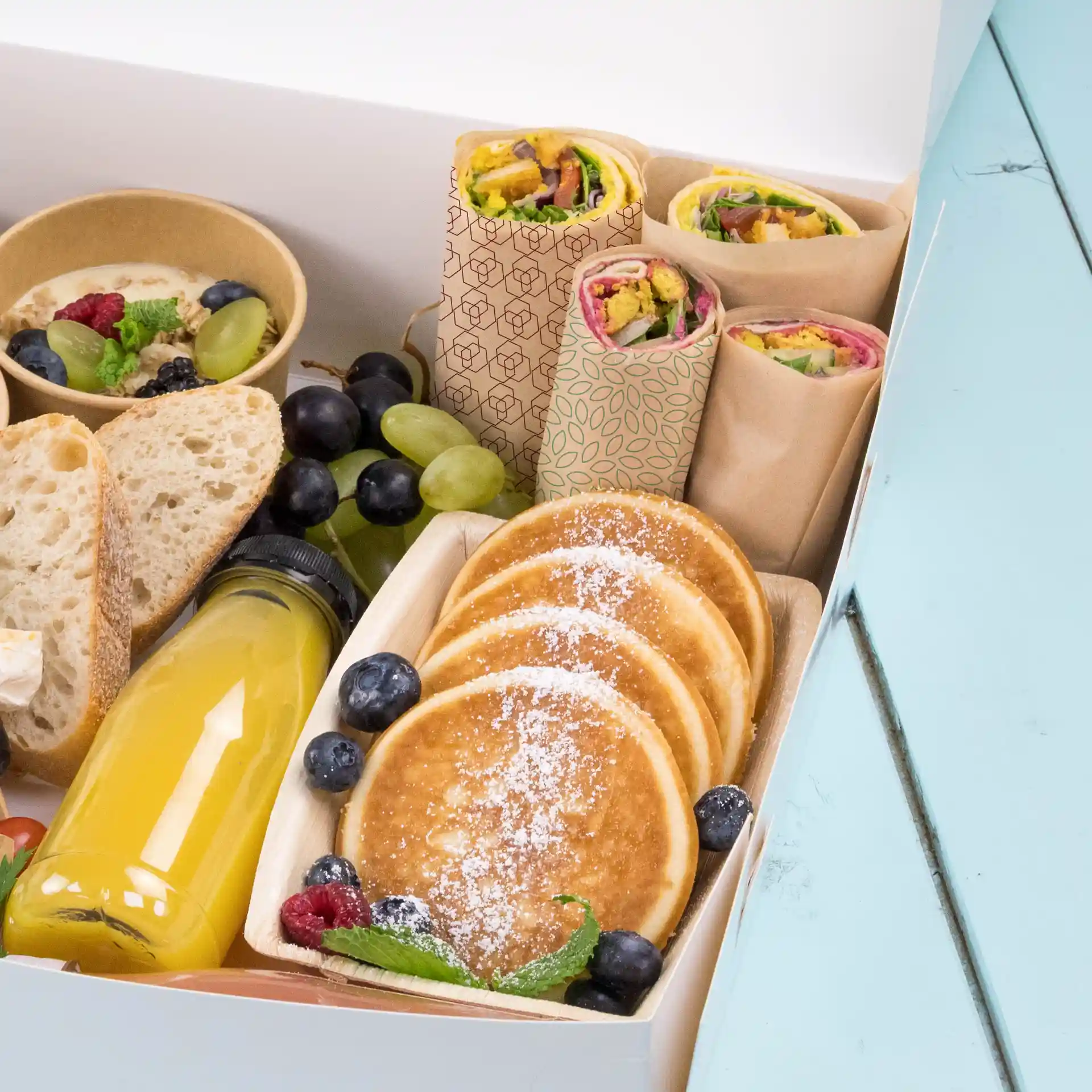 Assorted food items including wraps, pancakes, fruit, and a drink in a box on a blue surface.