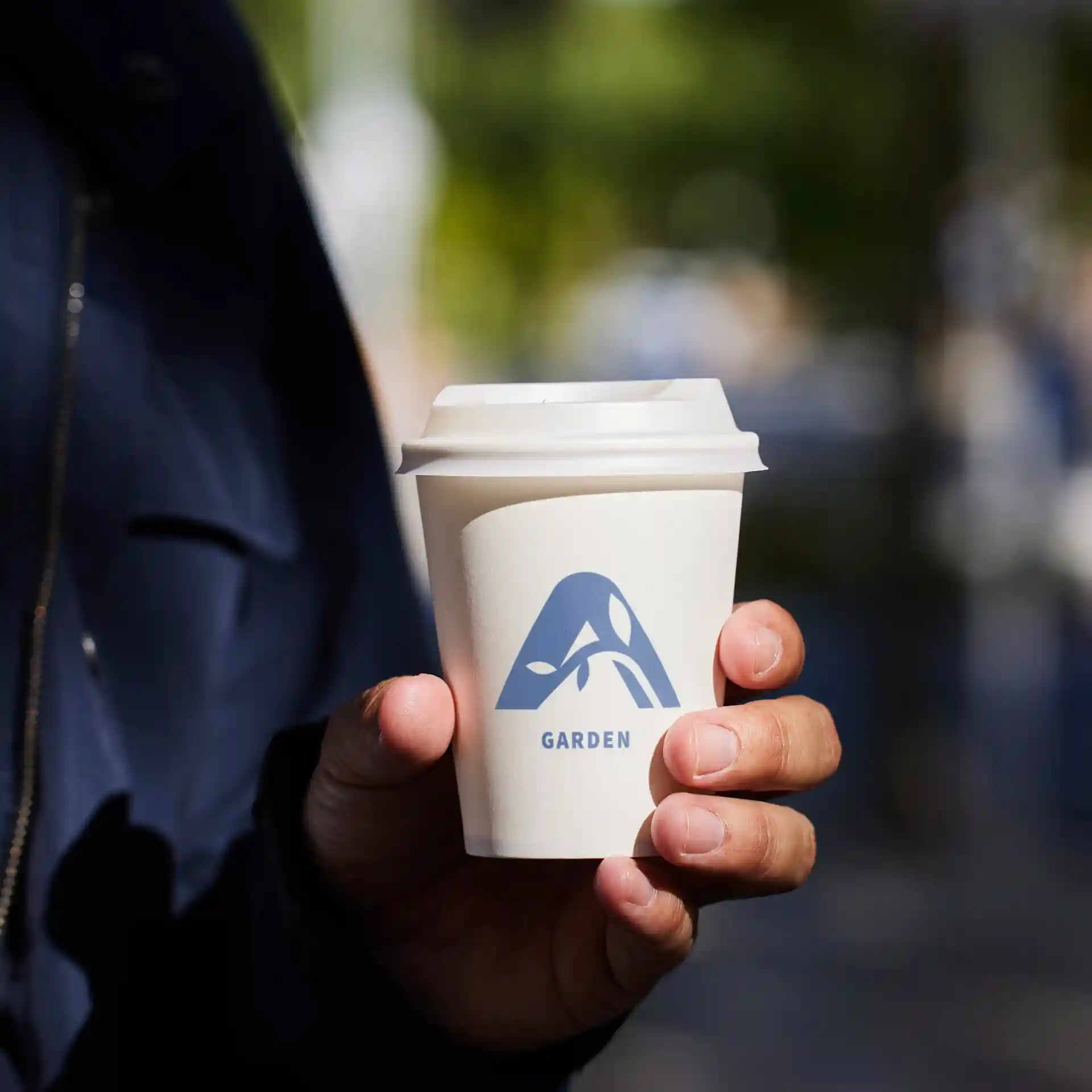 White paper cup with a blue logo and the word "GARDEN," held in a hand, outdoors with blurred background.