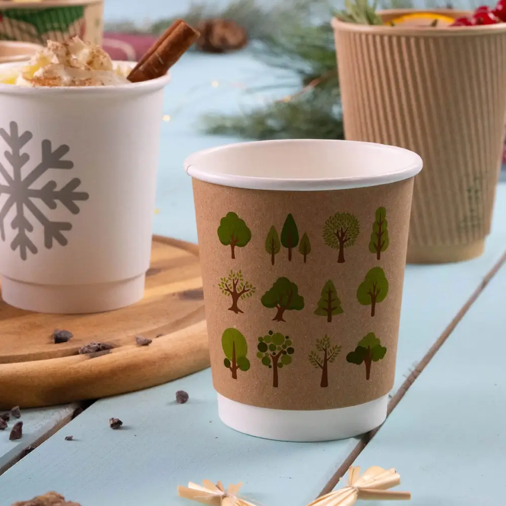 Kraft paper cup featuring green tree and plant graphics, placed on a wooden surface with other drink cups in the background.