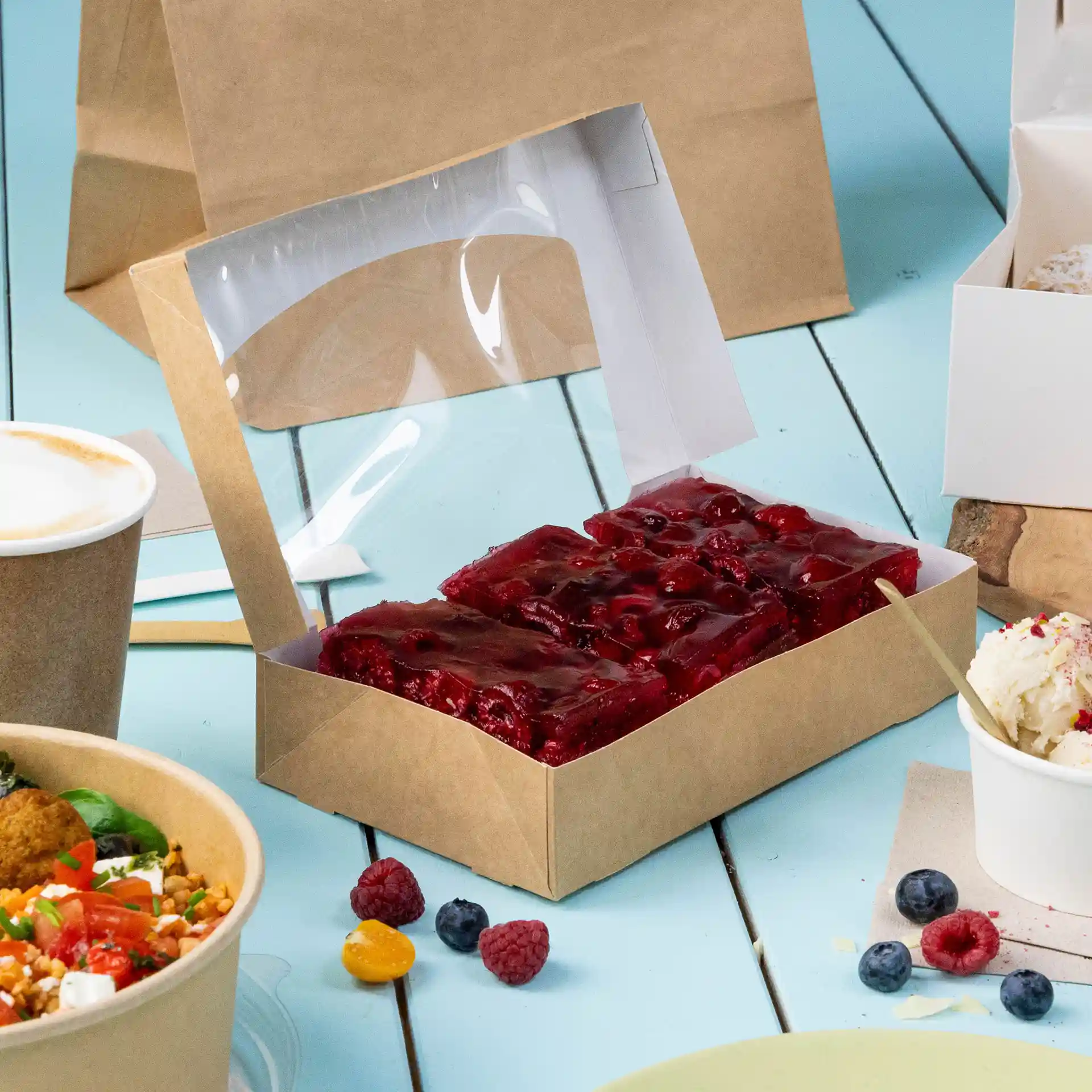 Cardboard window box with a transparent lid, containing red jelly dessert, on a blue surface with other food items.