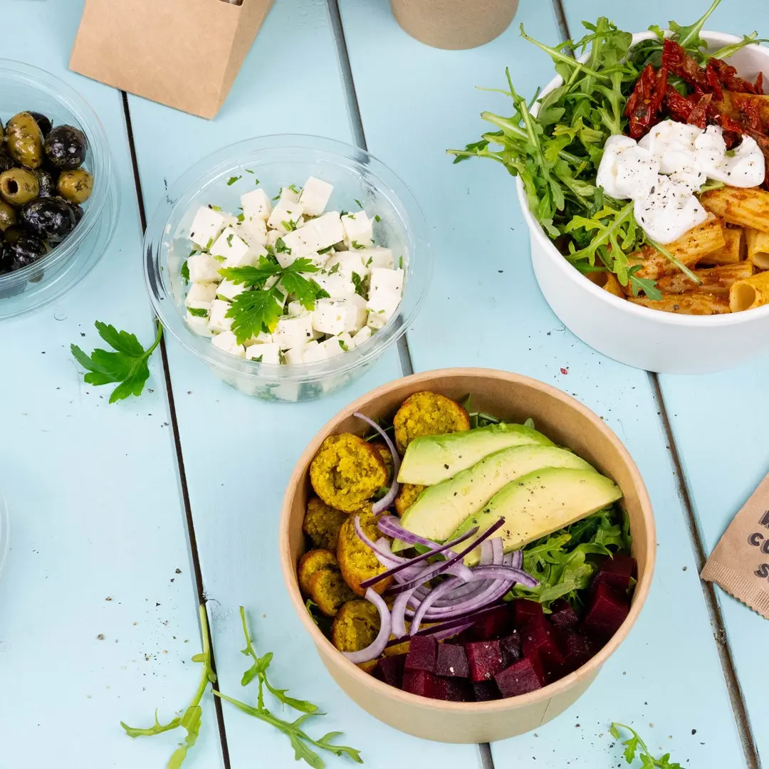 Bowl with falafel, avocado, beets, red onion, and greens, alongside containers of feta cheese and olives on a blue surface.
