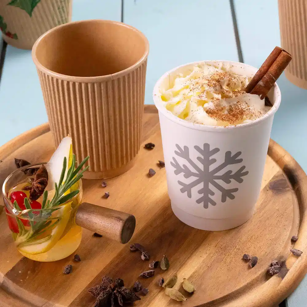 Image of a white double-walled paper cup with a snowflake design filled with whipped cream, beside a brown ribbed paper cup and a decorated glass.