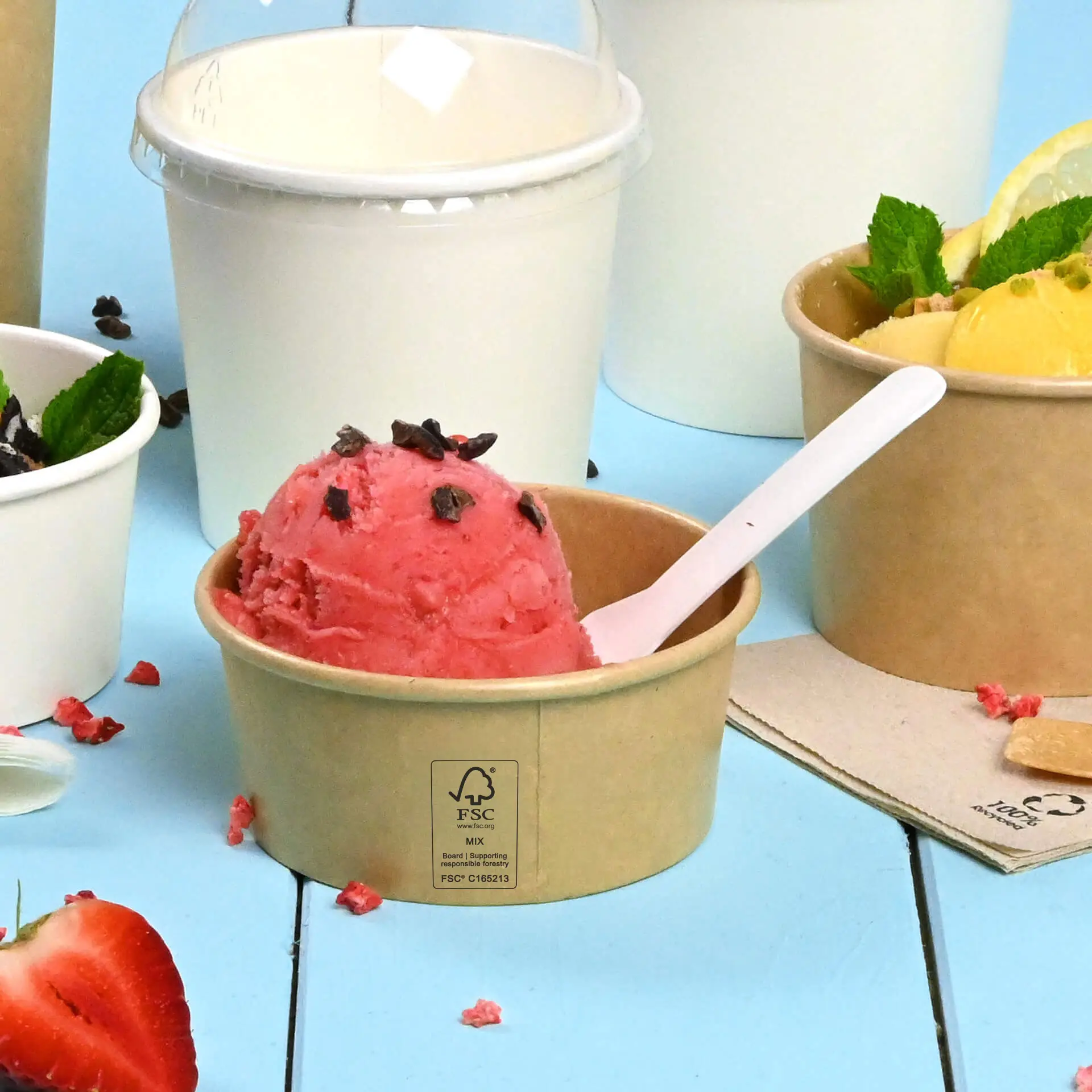 A brown, PE-coated ice cream cup with pink ice cream and chocolate sprinkles; other ice cream containers are partially visible.