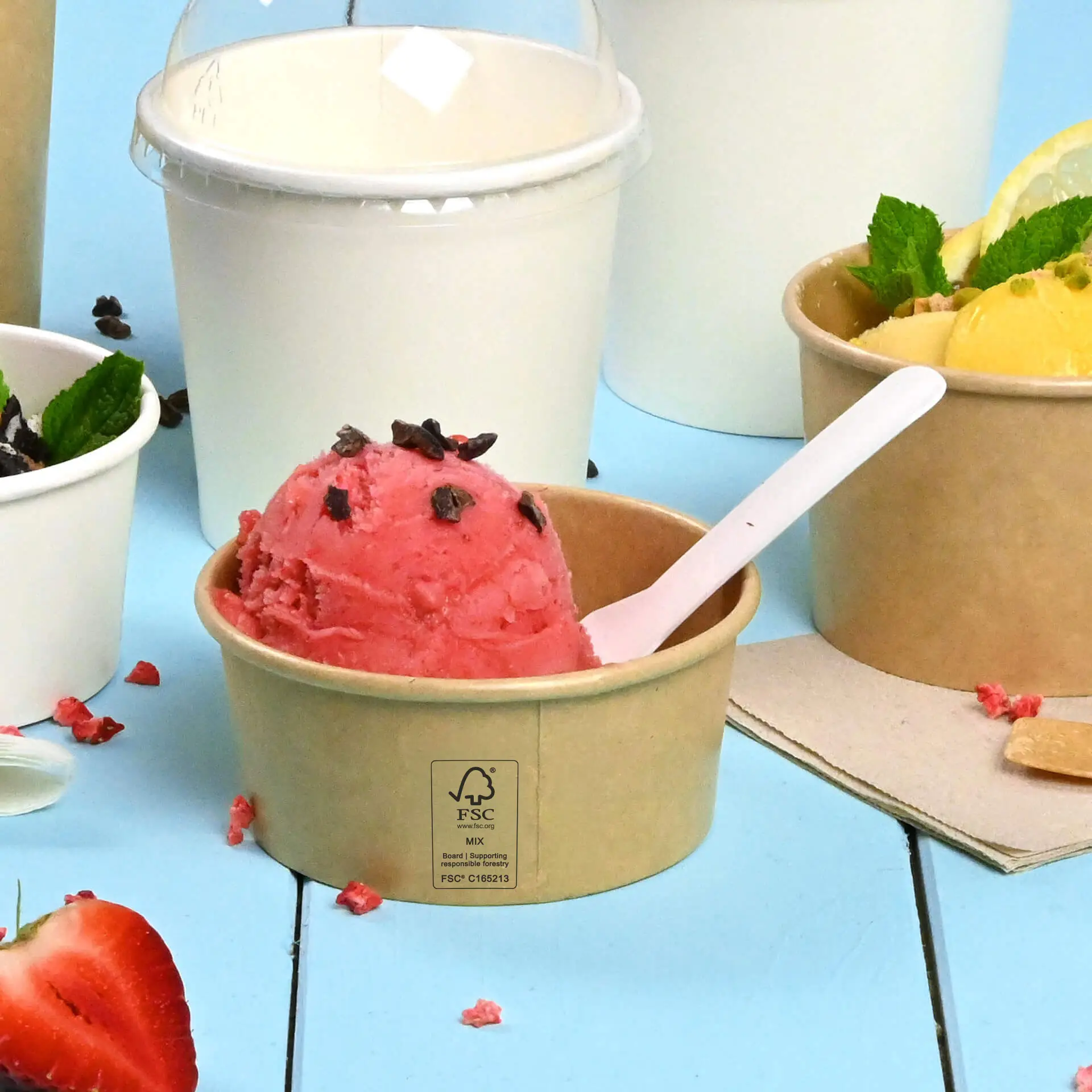 Brown paper cup with pink ice cream topped with chocolate chips, white spoon, and other cups in the background.