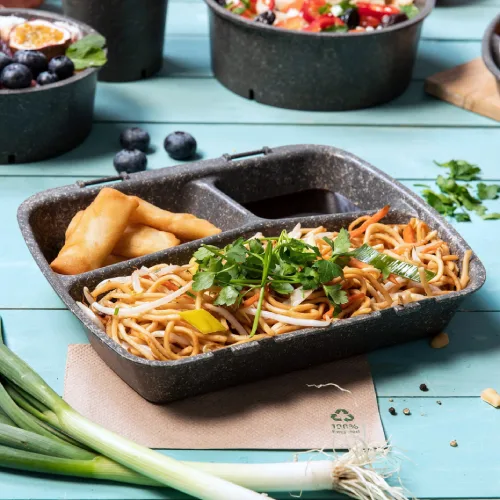 Meal box containing noodles, vegetable garnish, and spring rolls, placed on a blue wooden table with fresh ingredients around.