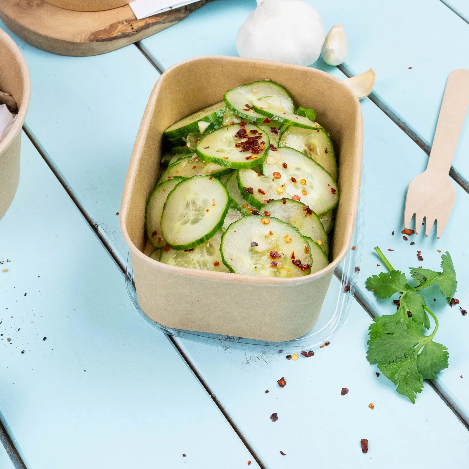 Brown kraft food container filled with sliced cucumbers, topped with chili flakes, placed on a light blue wooden surface.