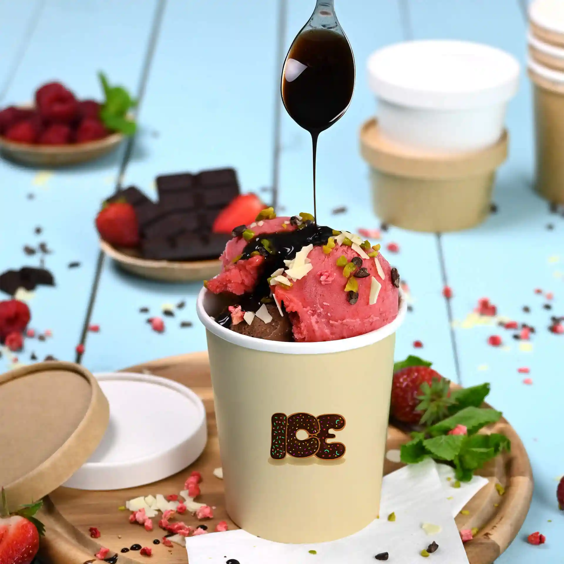 Round white ice cream cup with pink and brown scoops, topped with chocolate sauce and nuts, on a wooden tray.