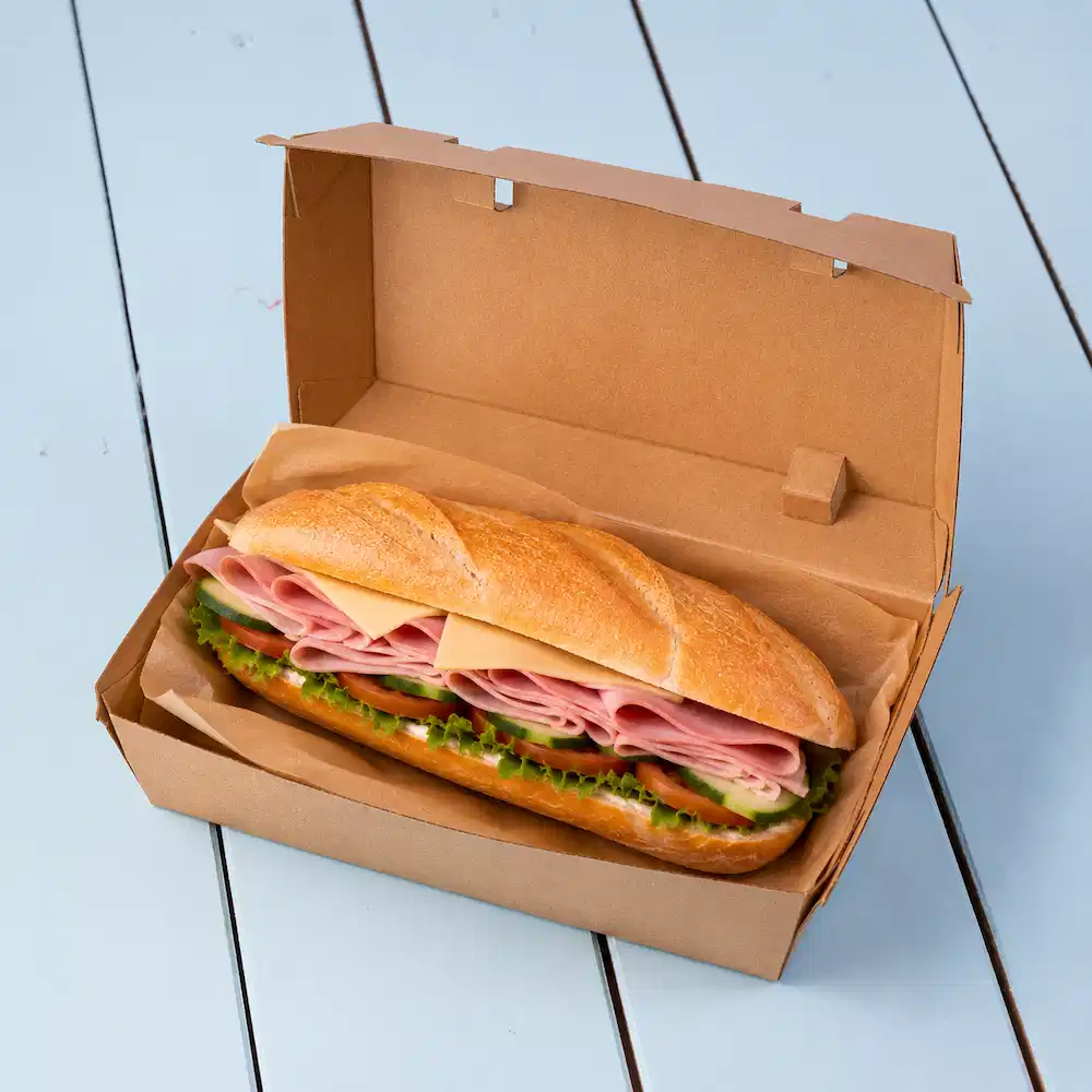 Sandwich in a brown takeout box, featuring layers of ham, cheese, lettuce, and sliced cucumbers.